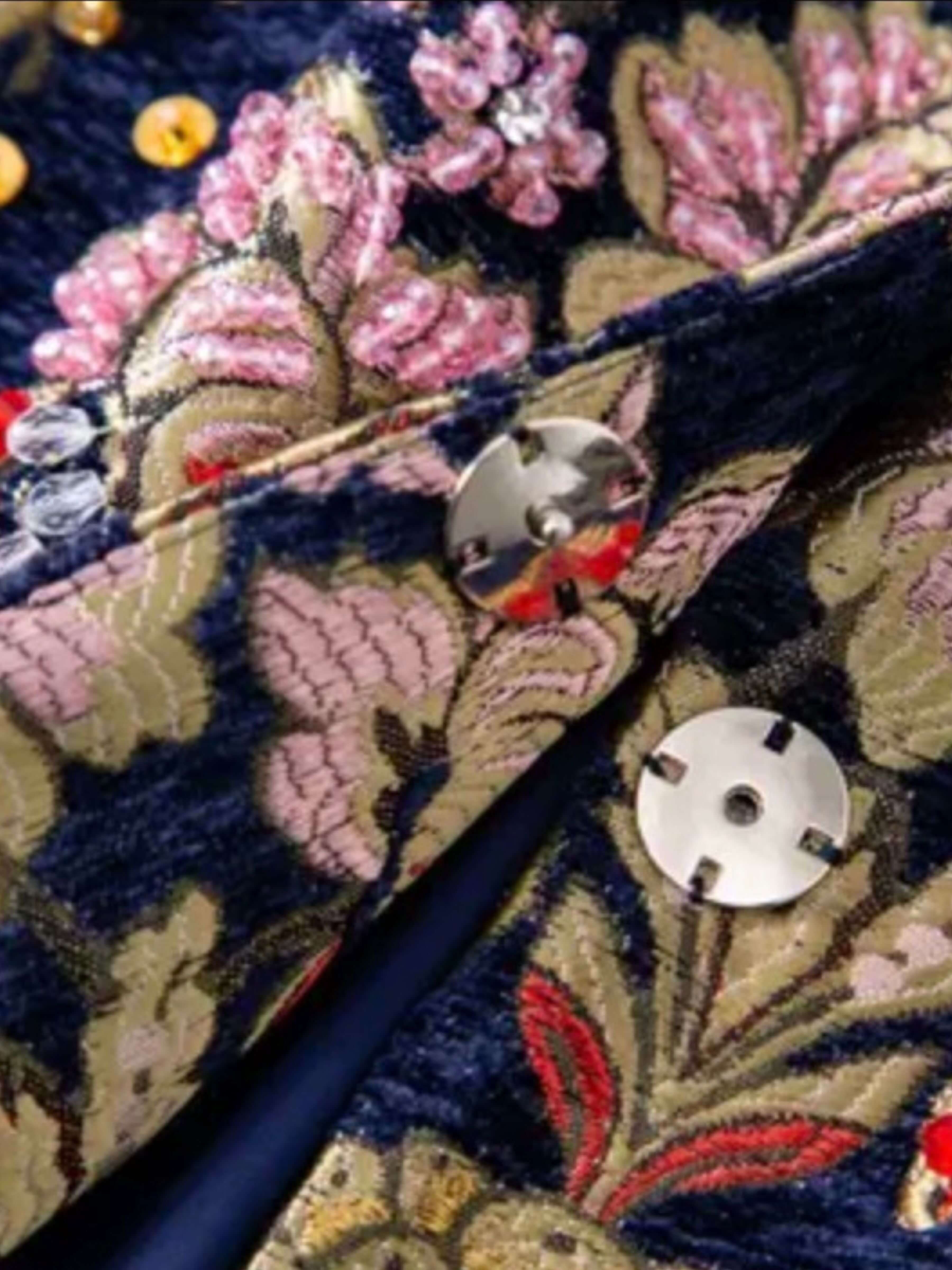 Close-up of a dark navy velvet jacket with an intricate floral brocade pattern in pink, gold, and red, featuring silver metal snap buttons.