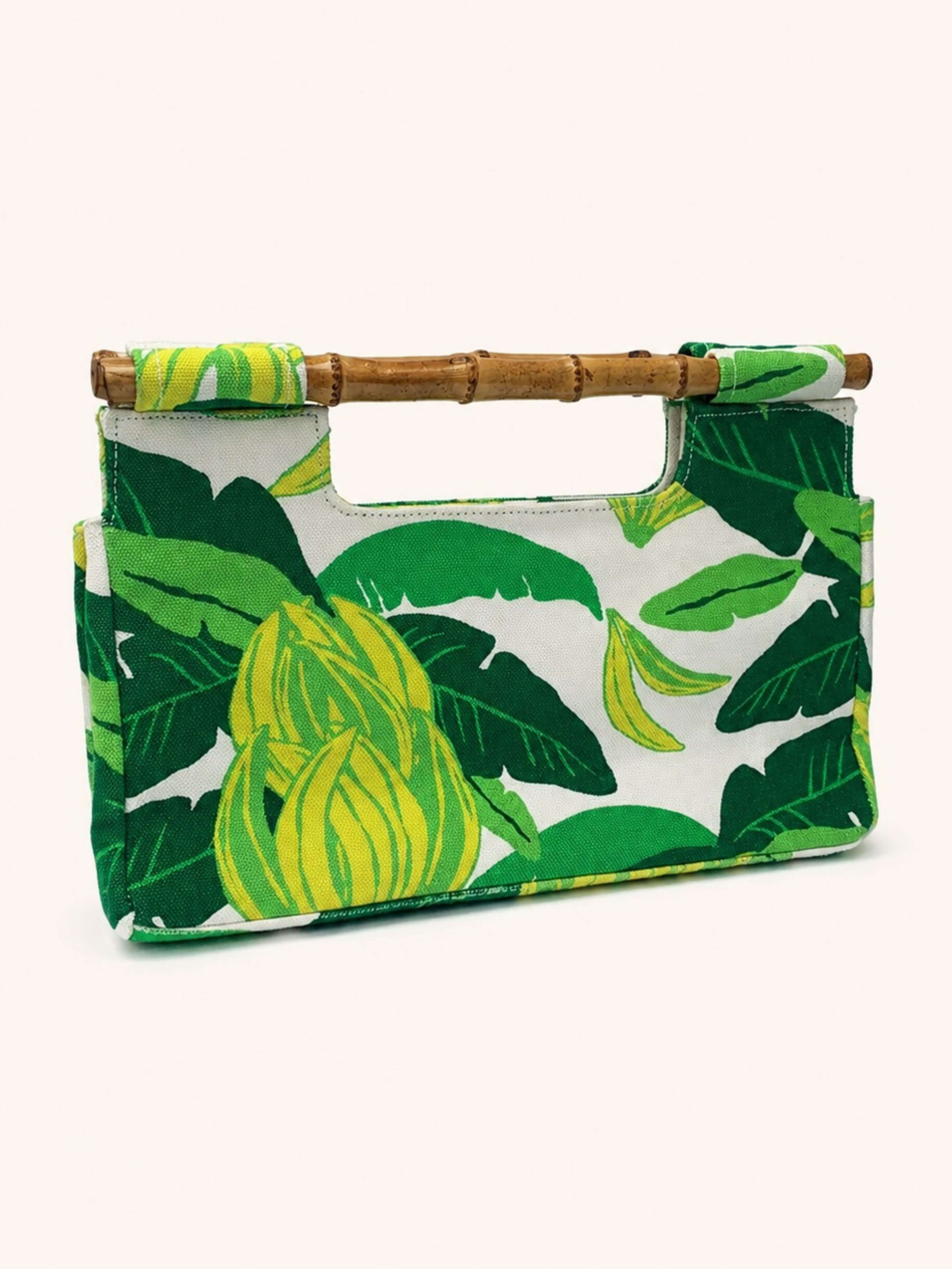 A rectangular handbag with a green and yellow banana leaf print on a white background, featuring a bamboo-style top handle, photographed against a solid off-white background.