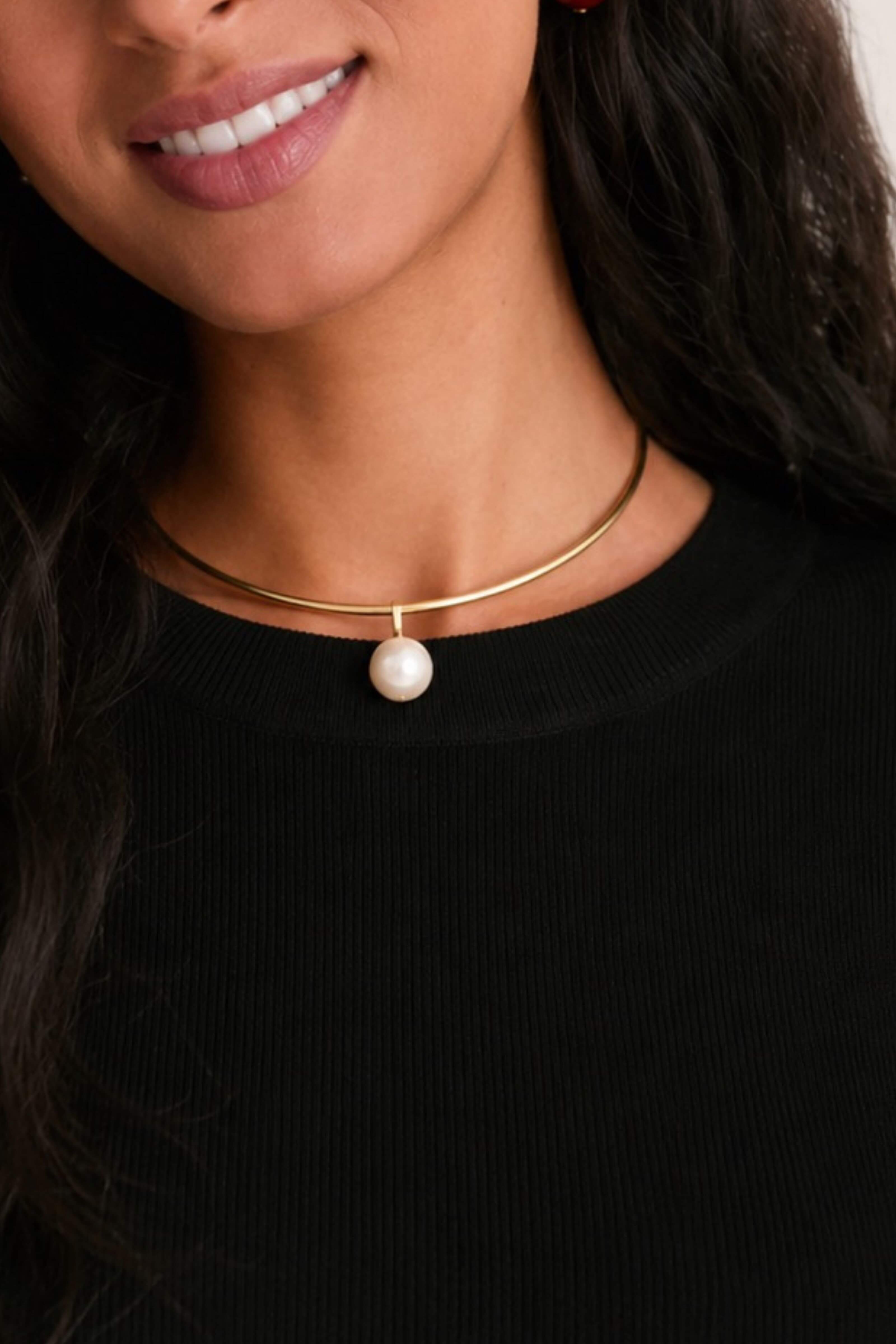 A close-up of a model wearing a delicate gold choker necklace with a single white pearl pendant, paired with a black ribbed top.
