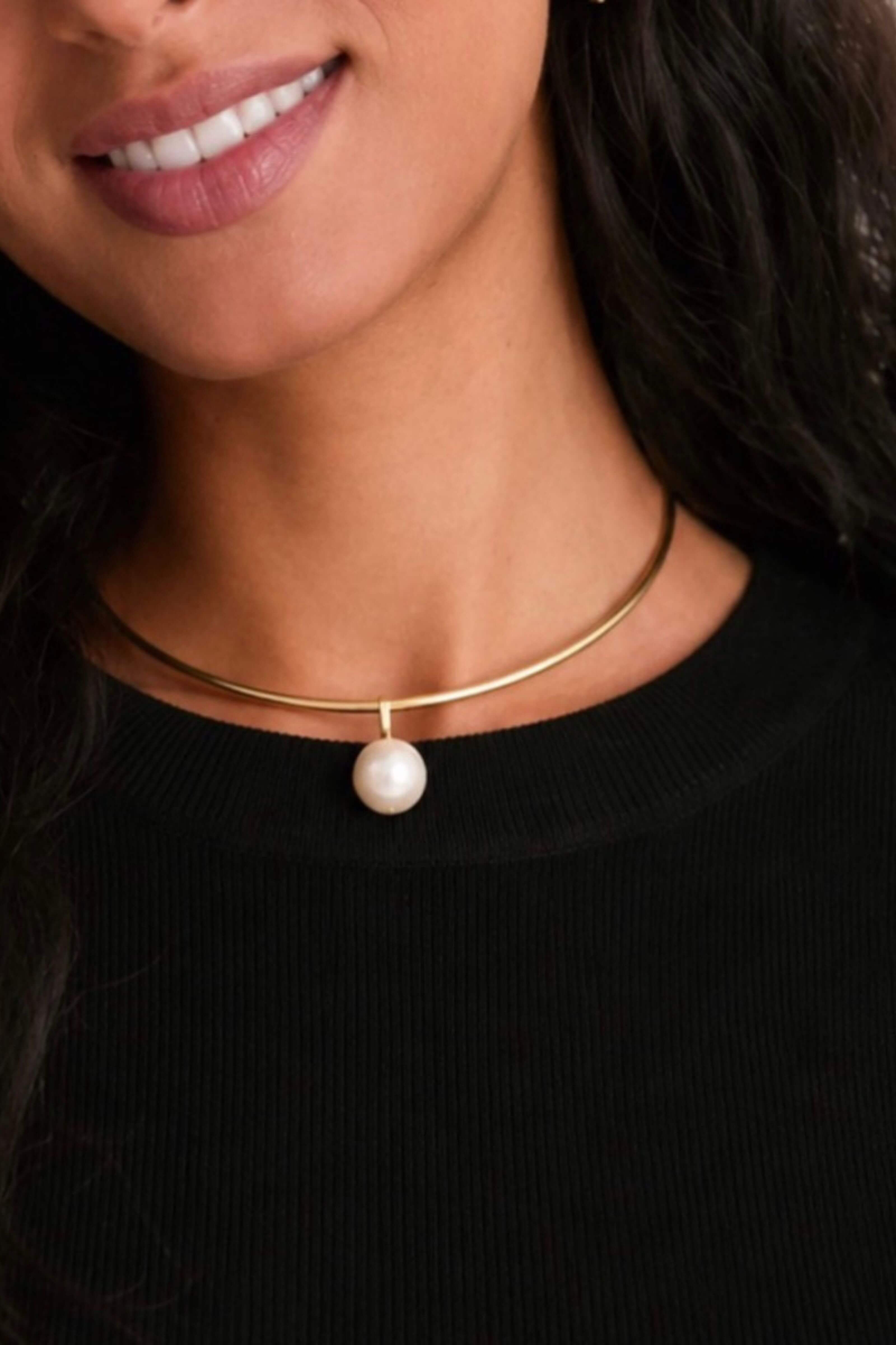 A close-up of a model wearing a delicate gold bangle necklace with a single white pearl pendant, paired with a black ribbed top.