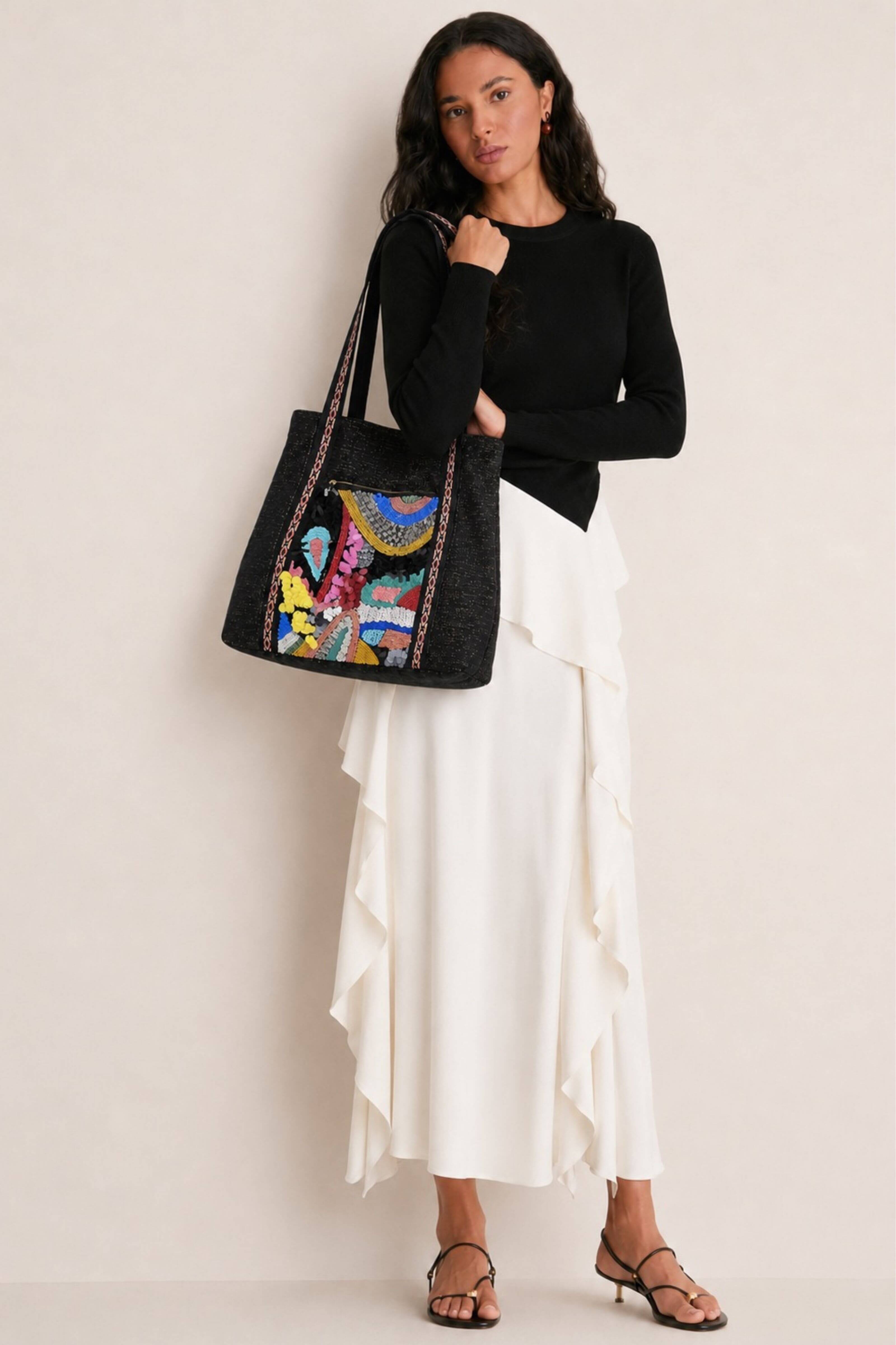 A woman modeling a black woven tote bag with a vibrant, multicolored embroidered design on the front, styled with a black long-sleeve top and a white ruffled midi skirt.