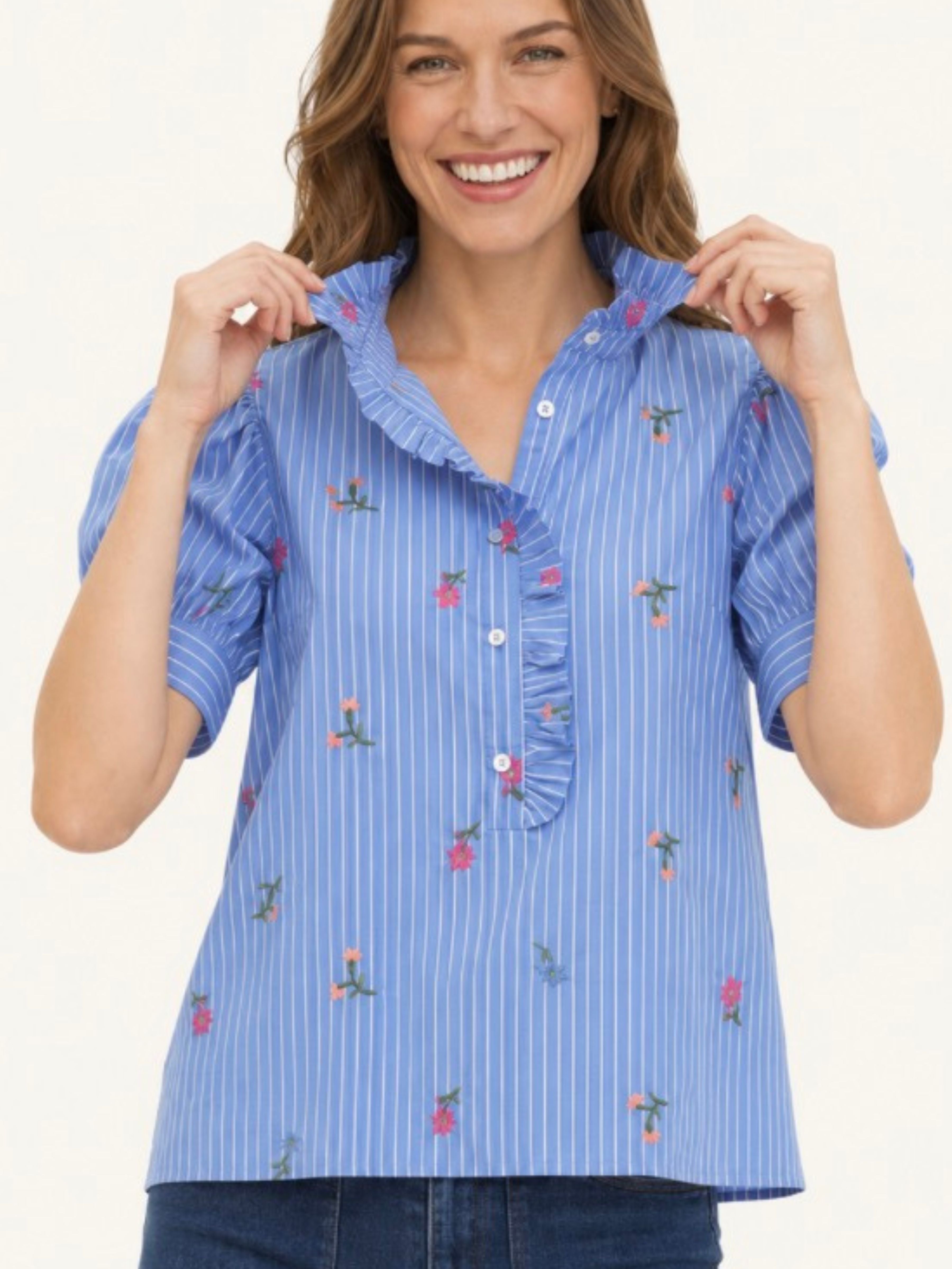 A woman wearing a light blue and white vertically striped short-sleeve blouse with ruffled trim and small pink floral embroidery, paired with dark blue jeans, against a plain white background.