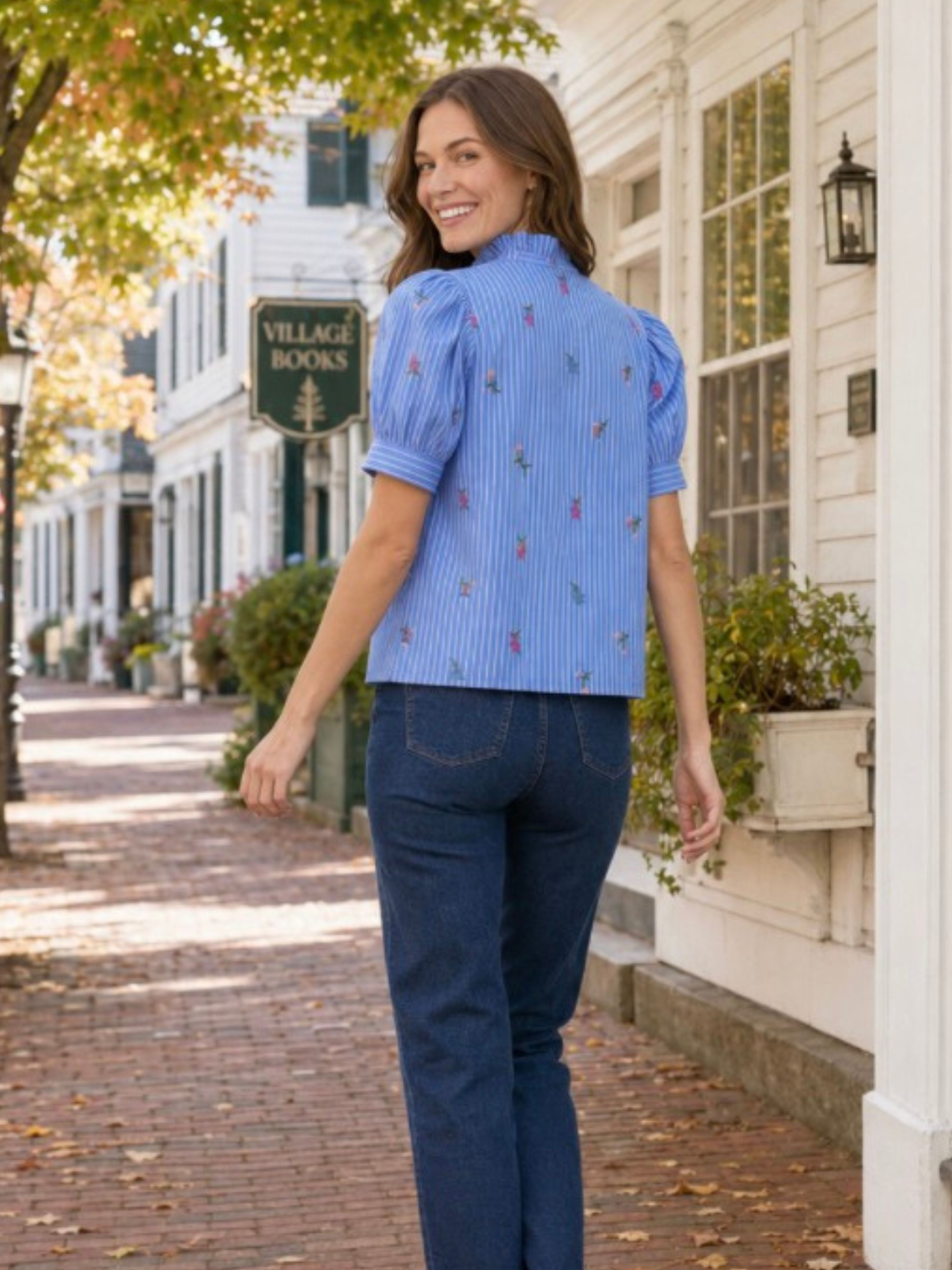 A woman wearing a light blue short-sleeve button-up shirt with vertical pinstripes and small colorful embroidered motifs, paired with dark blue jeans, walking down a brick sidewalk in front of a white building with a 'Village Books' sign.
