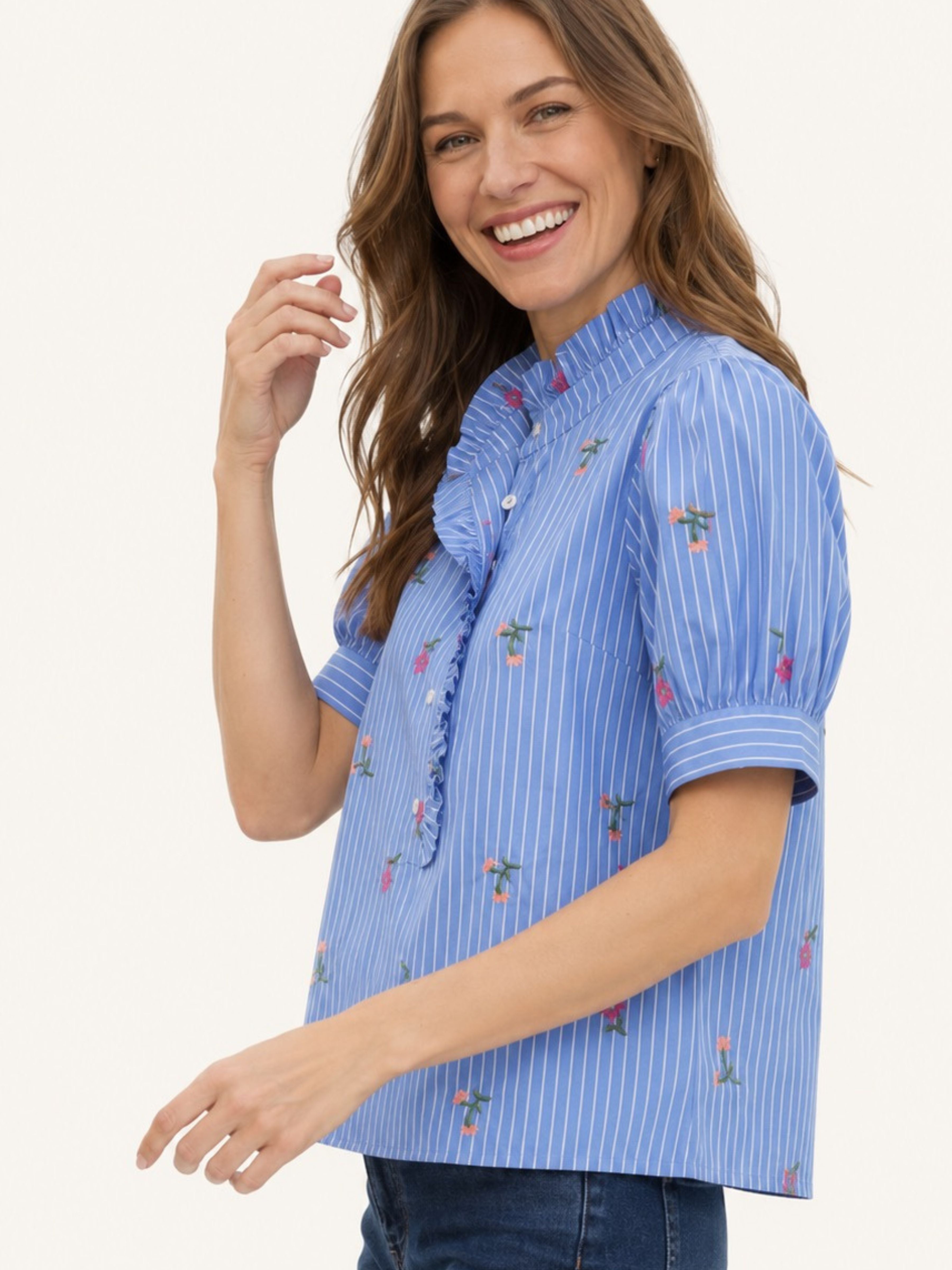 A woman wearing a light blue short-sleeve blouse with white vertical pinstripes and small embroidered floral details, paired with dark blue jeans, against a plain light background.