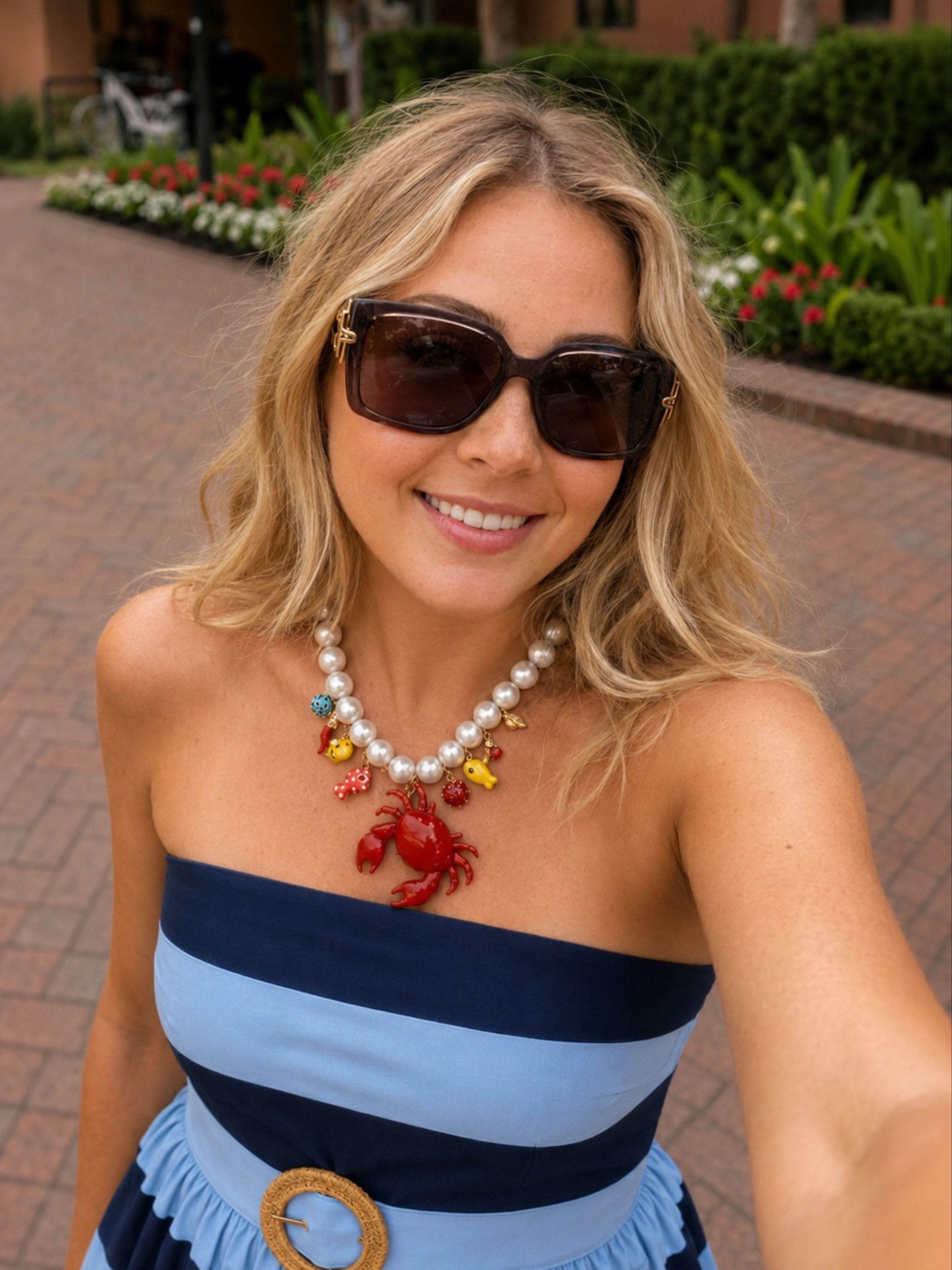 A woman wearing a blue and navy strapless dress with a light blue ruffled hem, accessorized with a pearl necklace featuring a red lobster charm, and dark sunglasses, posing outdoors on a brick walkway.
