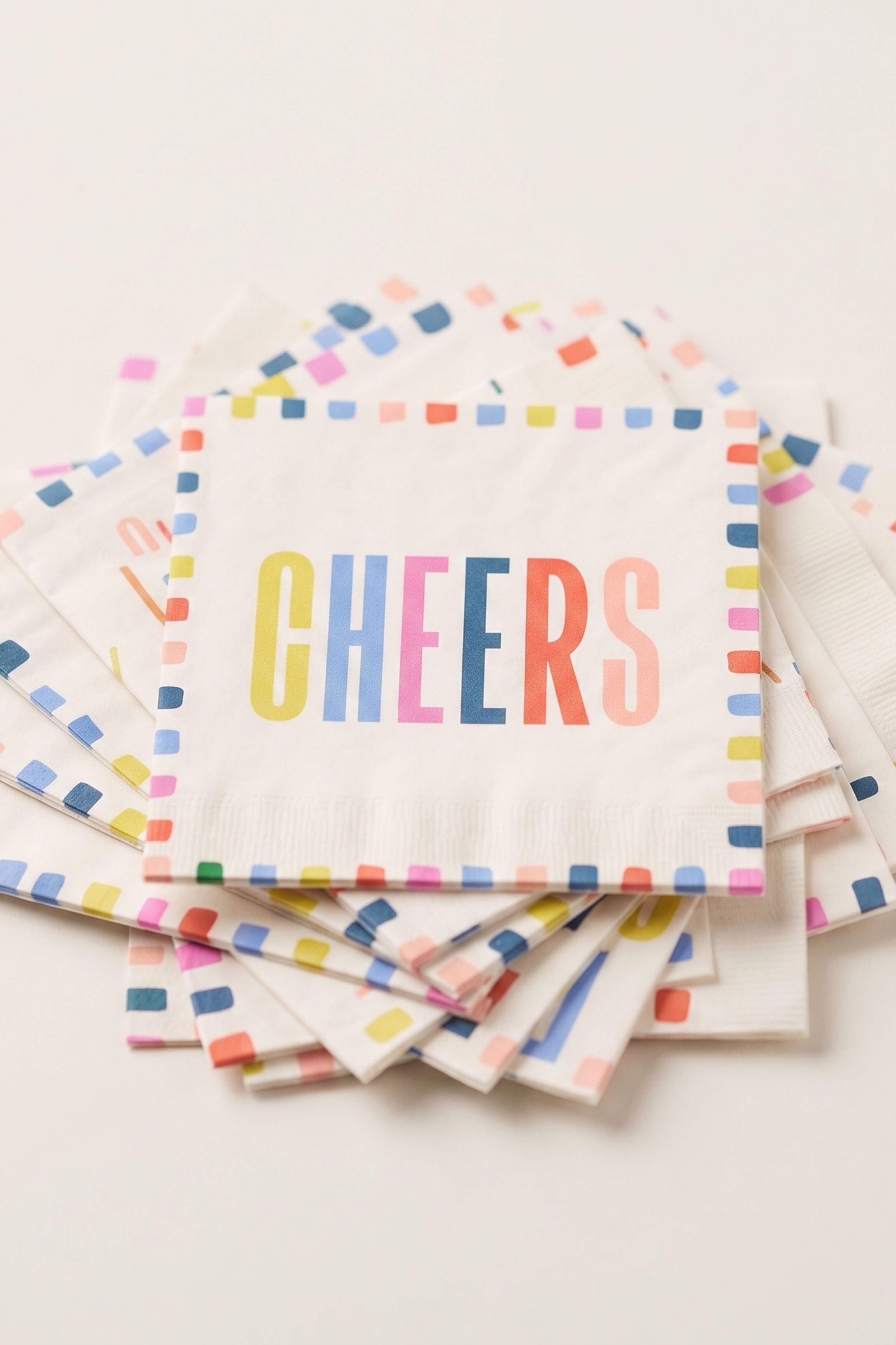 A stack of white paper party napkins with colorful confetti borders and the word 'CHEERS' printed in multicolored letters on the top napkin, photographed on a light neutral background.
