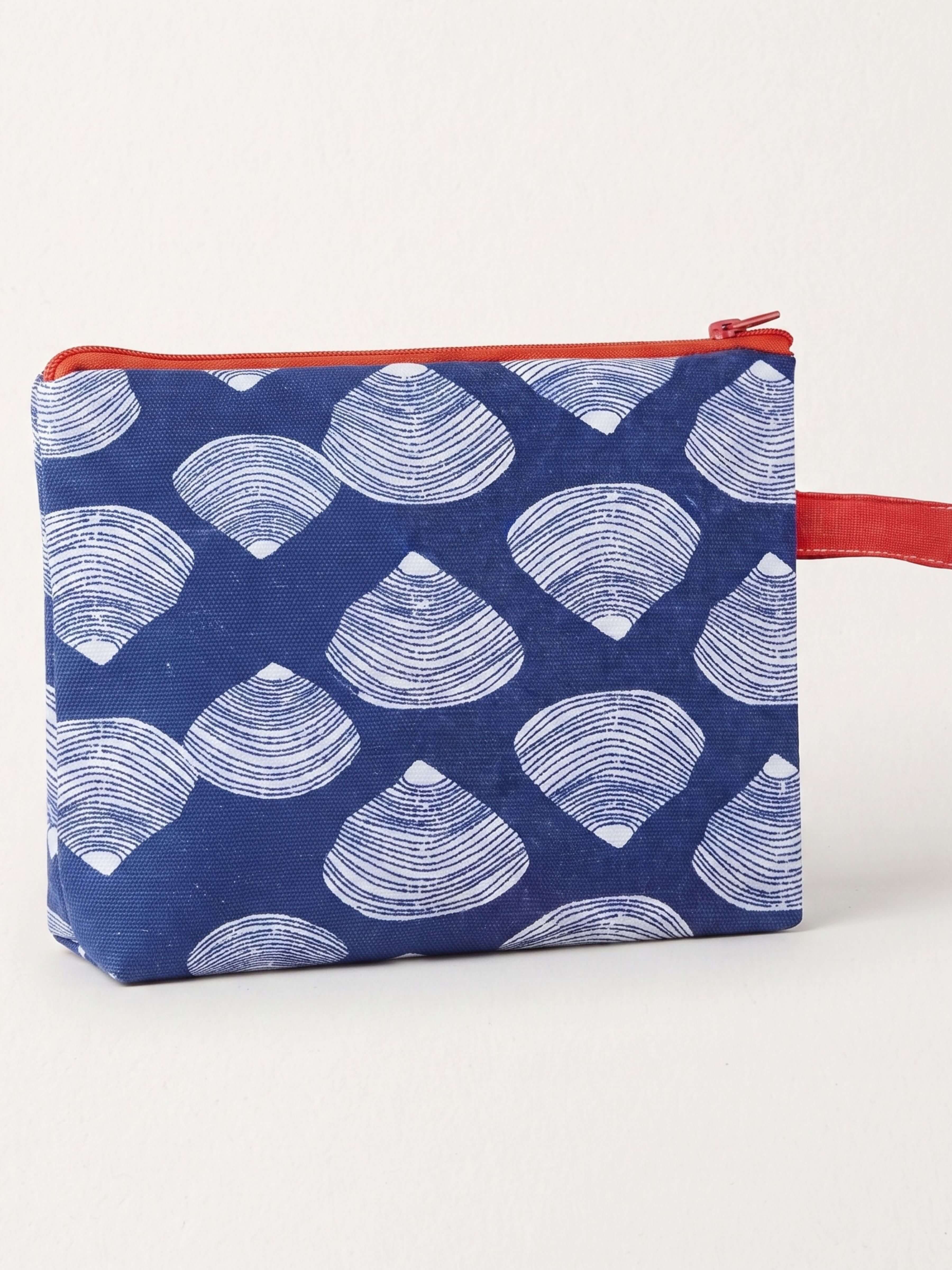 A small zippered pouch with a navy blue background and a repeating white scallop shell pattern, featuring a red zipper and a red wrist strap, shown on a light off-white background.
