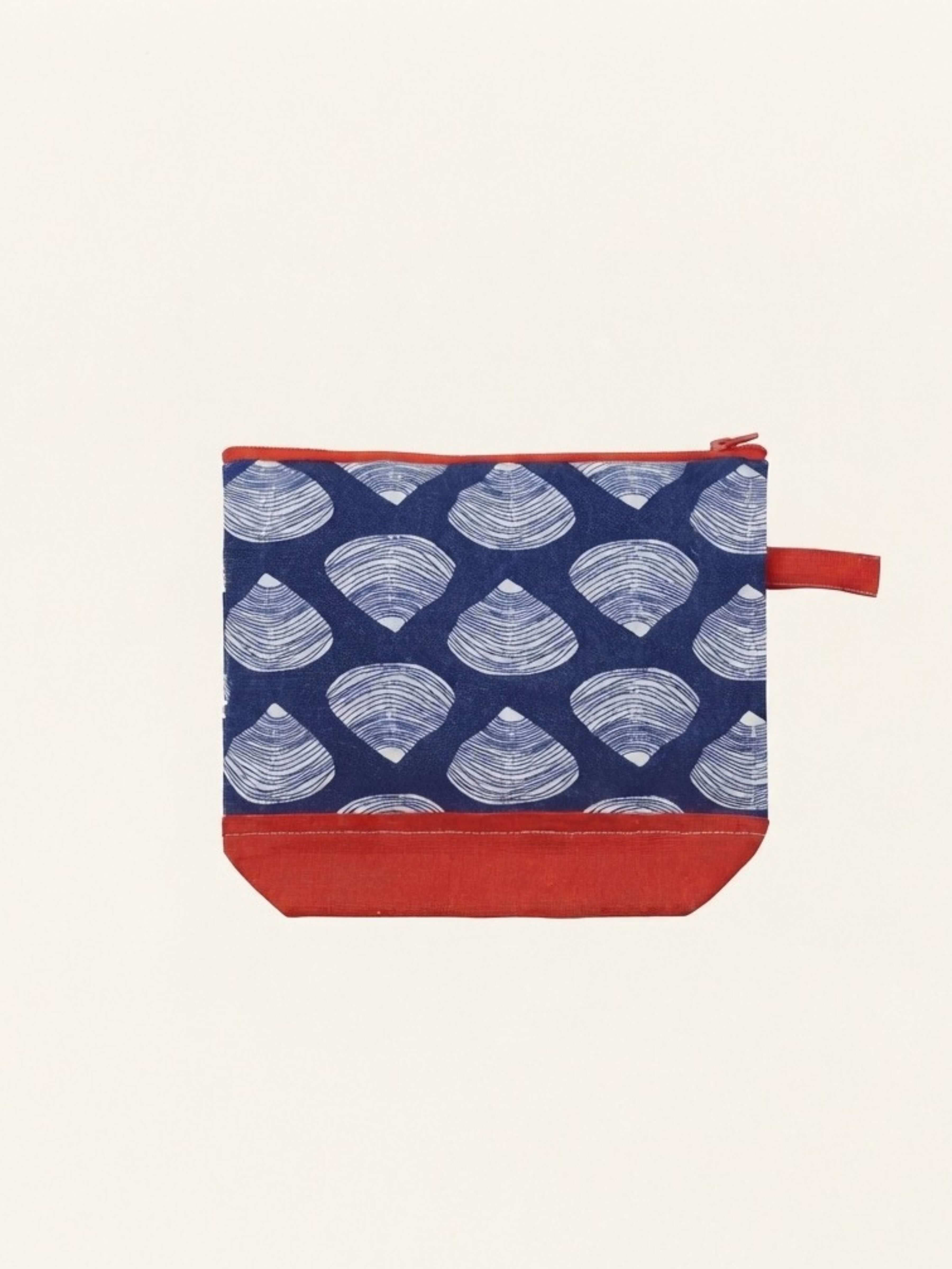 A small fabric zip pouch with a navy blue background, a repeating white scallop shell pattern, and solid red trim along the zipper, base, and carrying loop, shown on a plain off-white background.