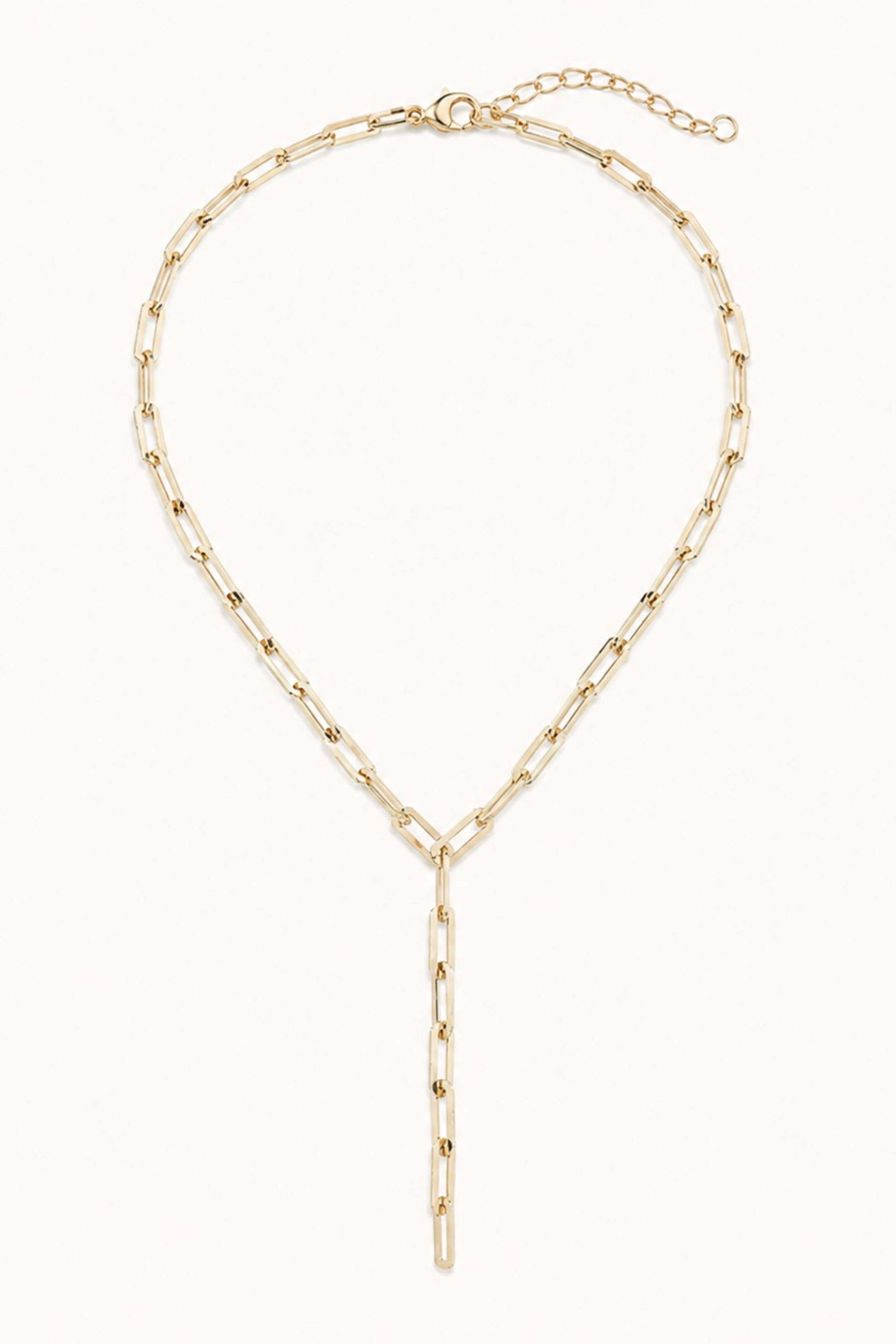 A gold paperclip chain necklace with a long vertical dangle pendant, displayed on a clean off-white background.