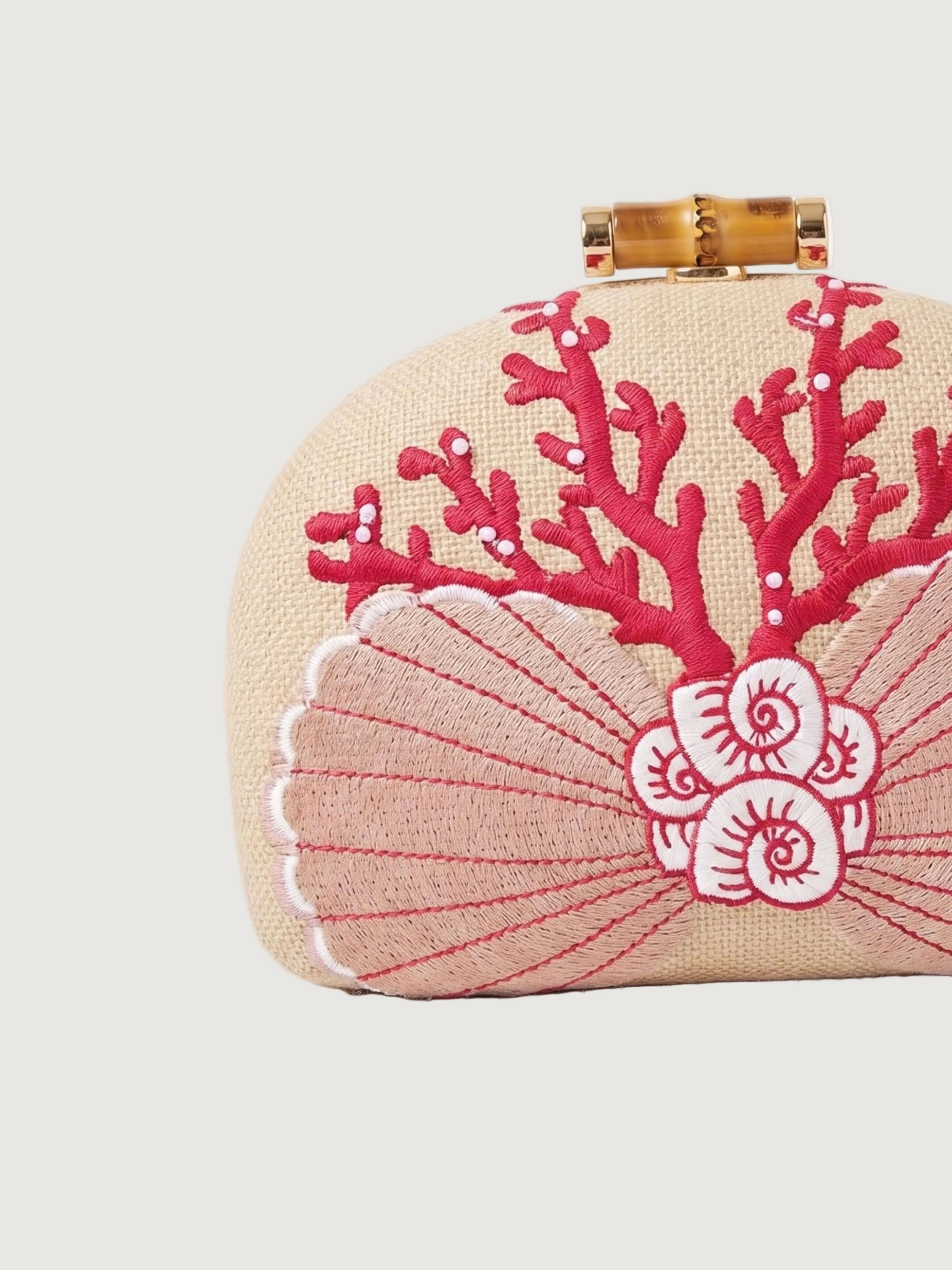 A close-up of a beige woven clutch bag with a bamboo-style clasp and embroidered coral and shell motifs in pink and white.
