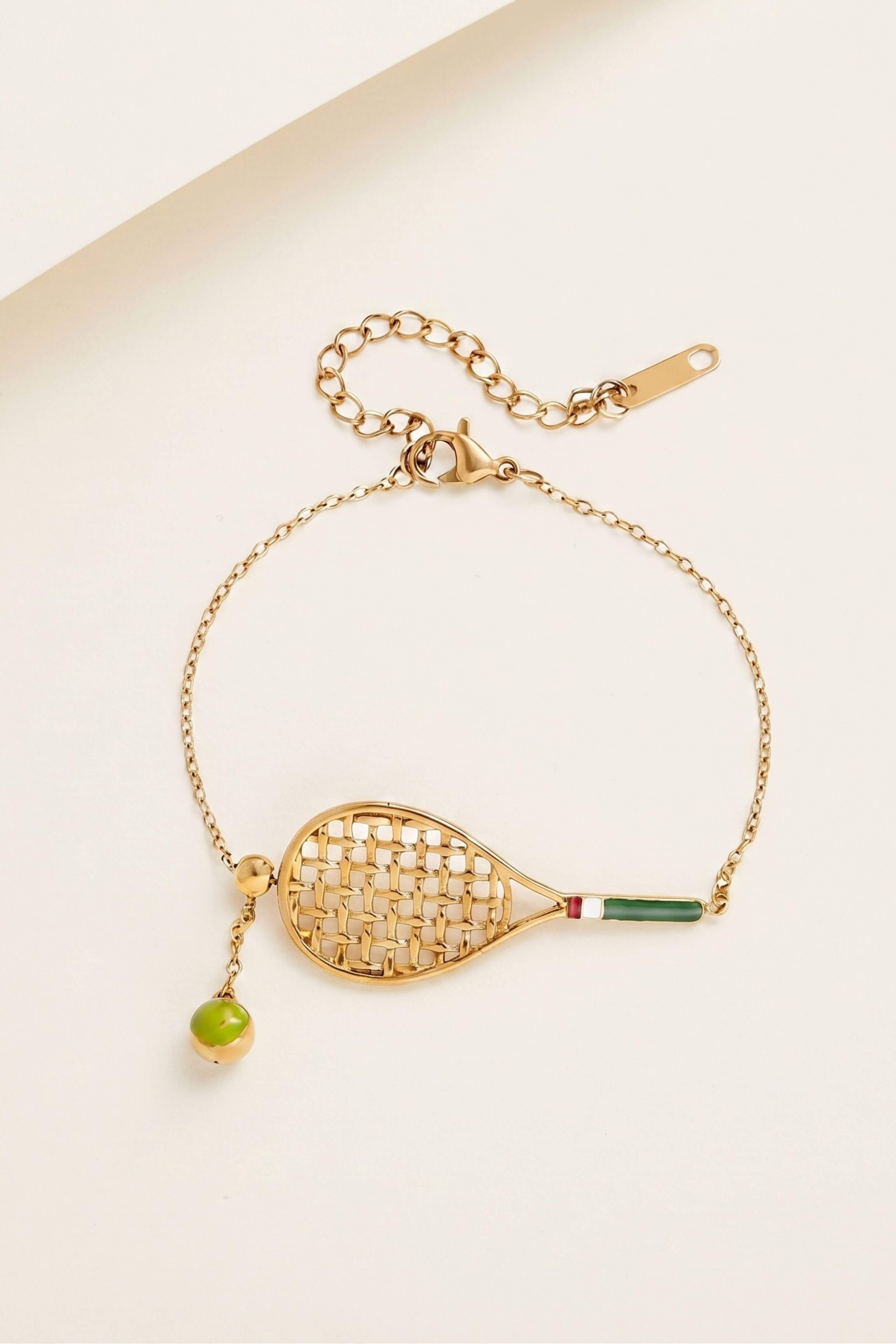A gold chain bracelet featuring a tennis racket pendant with a woven-texture center, a green and red handle, and a dangling green bead, displayed on a light beige background.