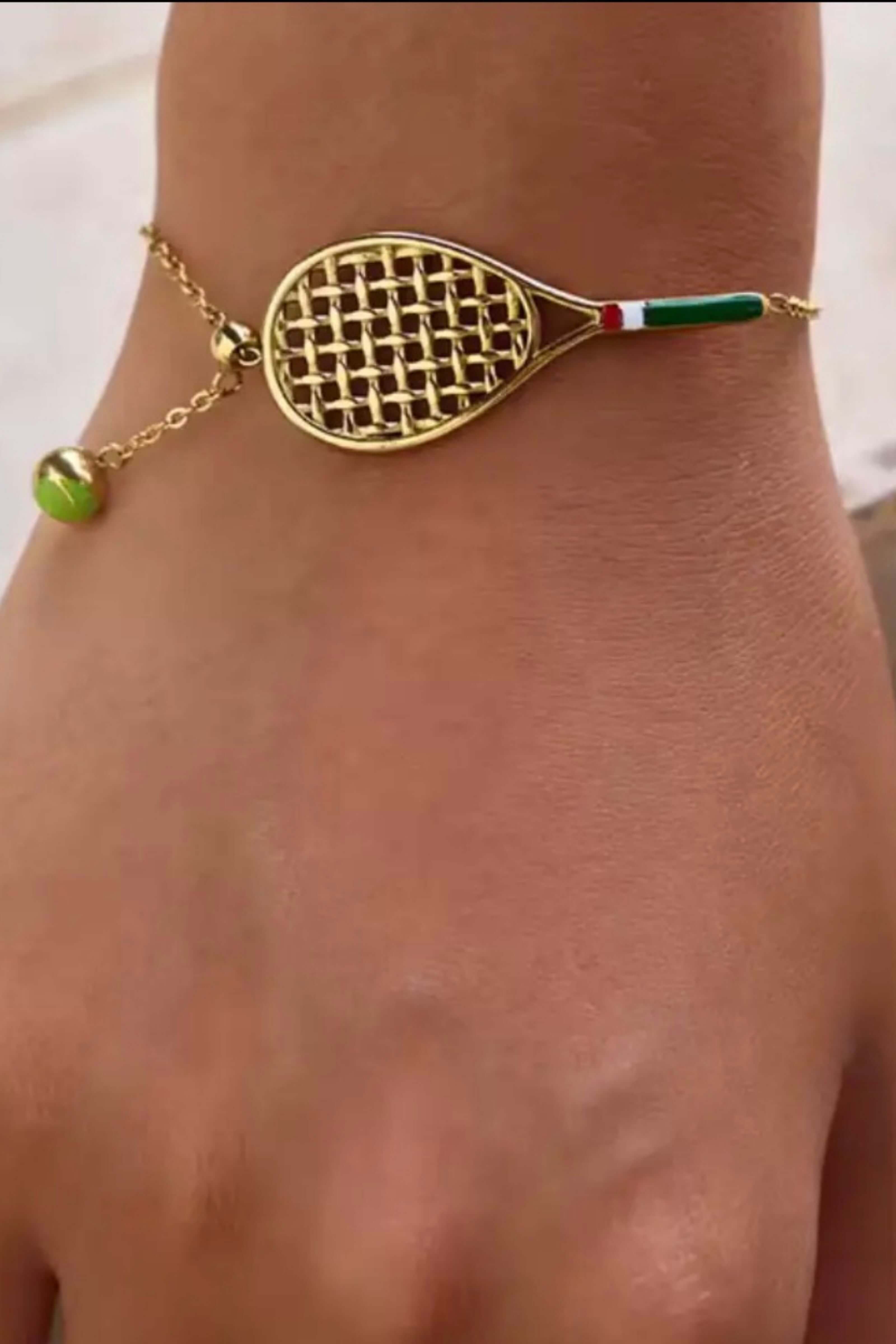 A gold-toned tennis bracelet shaped like a tennis racket, worn on a wrist. The bracelet has a gold mesh pattern on the racket head, a thin gold chain with a small green bead dangling on one side, and a handle featuring the green, white, and red colors of the Italian flag.