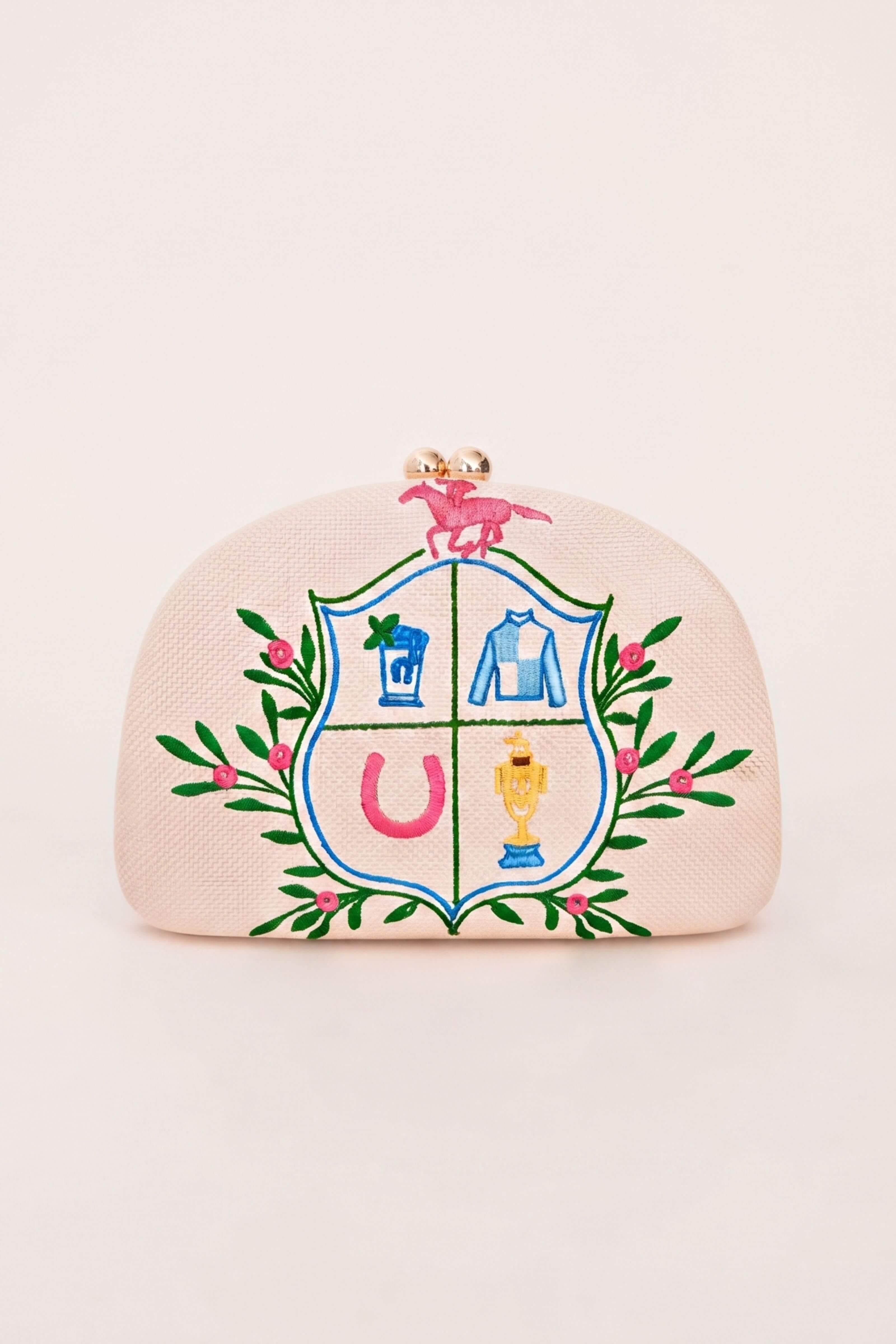 A light pink textured clutch purse with a gold-tone clasp and a colorful embroidered crest featuring horse, jacket, trophy, and horseshoe motifs, set against a solid cream background.