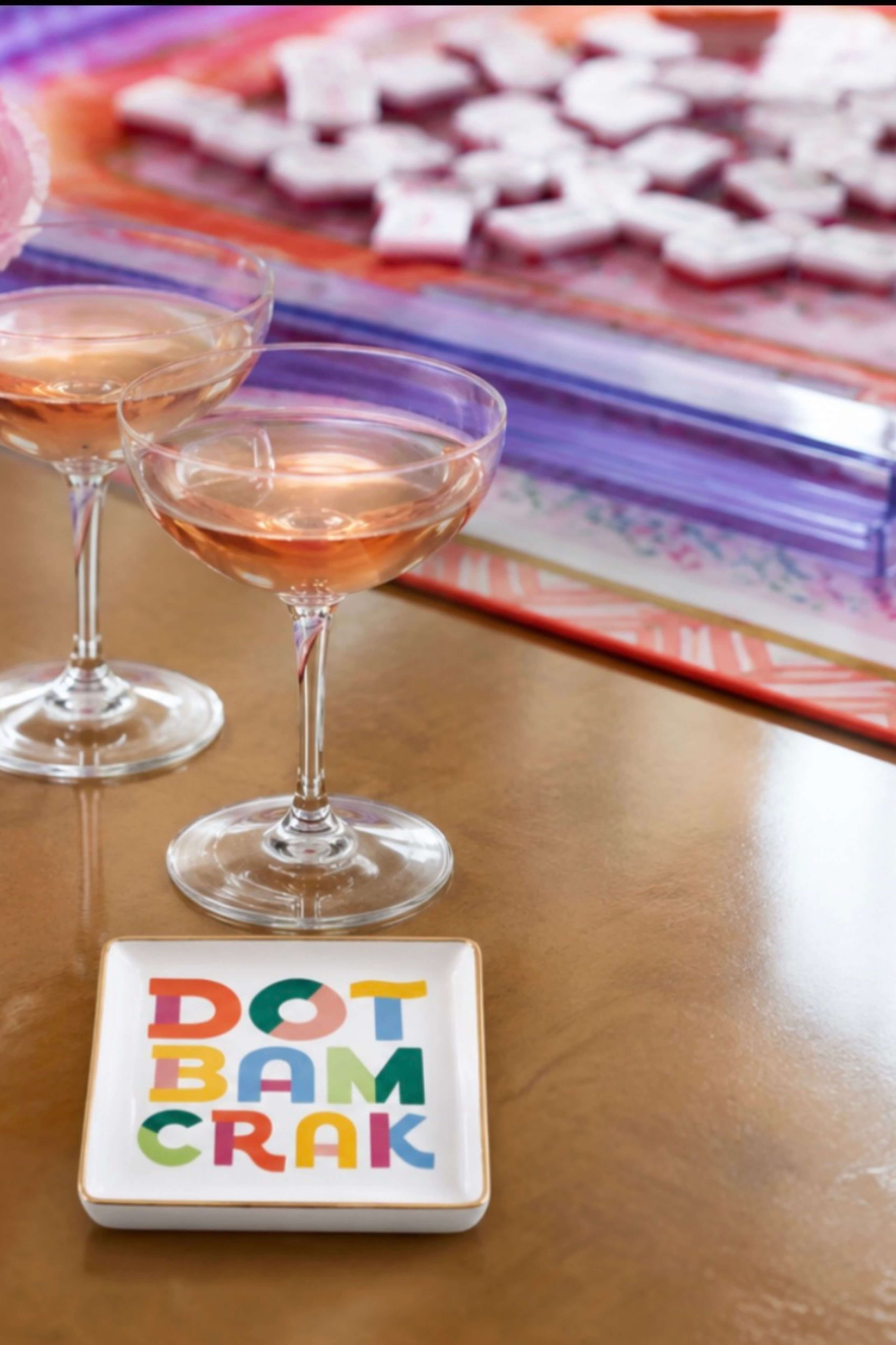 A square white coaster with the phrase 'DOT BAM CRAK' in colorful block letters sits on a wooden table next to two coupe glasses filled with pinkish beverage, with a blurred colorful tray in the background.
