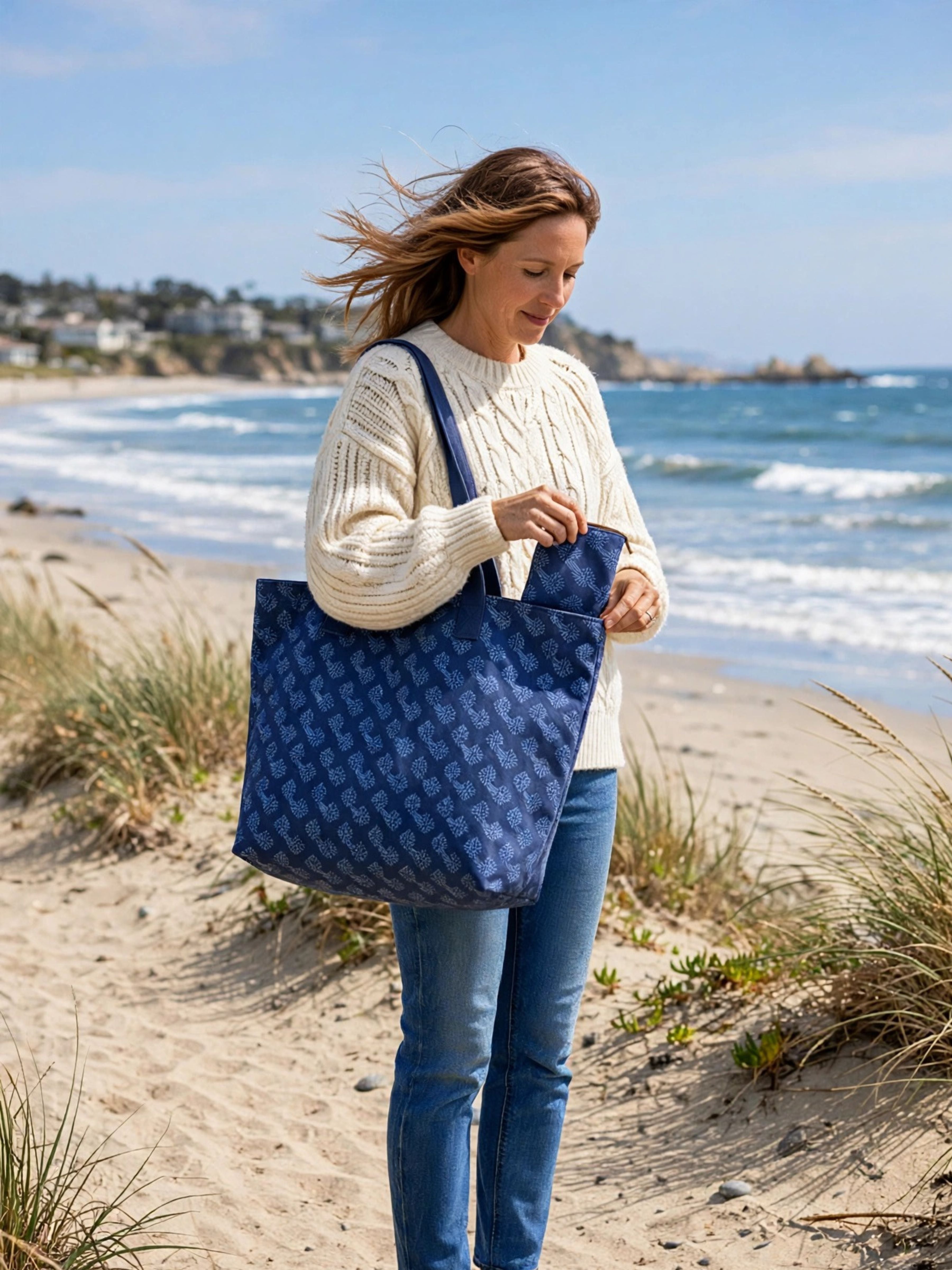 A woman standing on a sandy beach wearing a cream cable-knit sweater and blue jeans, carrying a large navy blue tote bag with a repeating tonal monogram pattern.