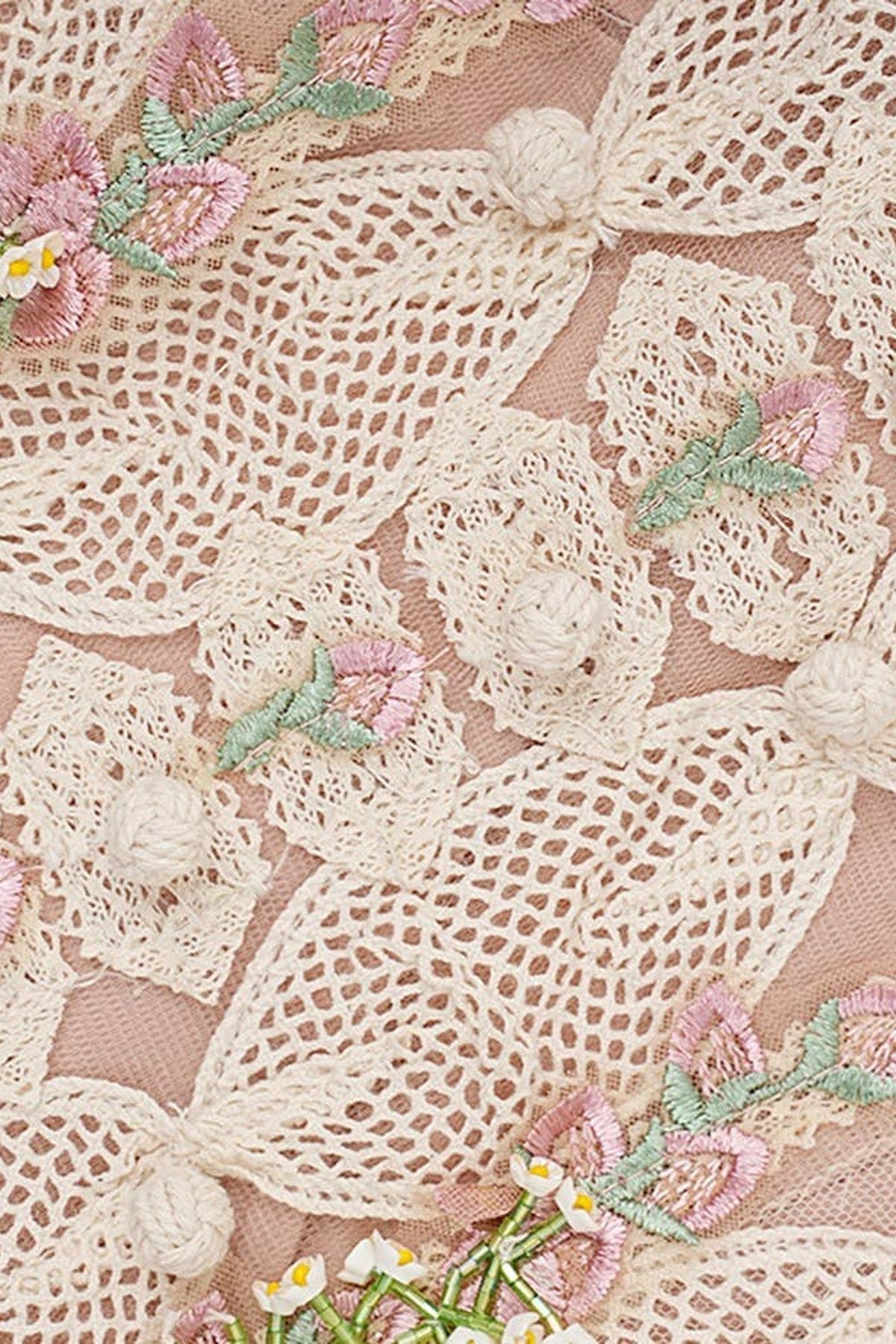 Close-up of a delicate cream lace fabric with embroidered pink flowers, green leaves, and small white pom-pom accents on a soft blush pink background.