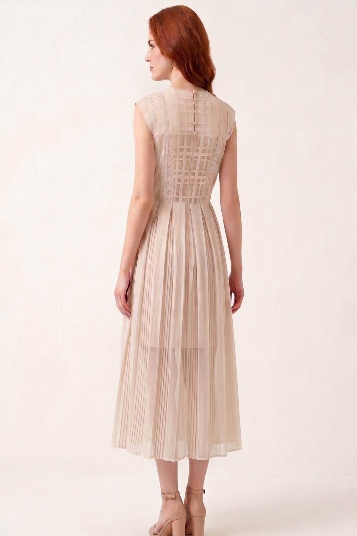 A woman with red hair is shown from the back wearing a sleeveless, pleated midi dress in a light beige or champagne color. The dress features a structured, crisscross lace-like pattern across the upper back and a pleated skirt.