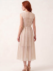 A woman with red hair is shown from the back wearing a sleeveless, pleated midi dress in a light beige or champagne color. The dress features a structured, crisscross lace-like pattern across the upper back and a pleated skirt.