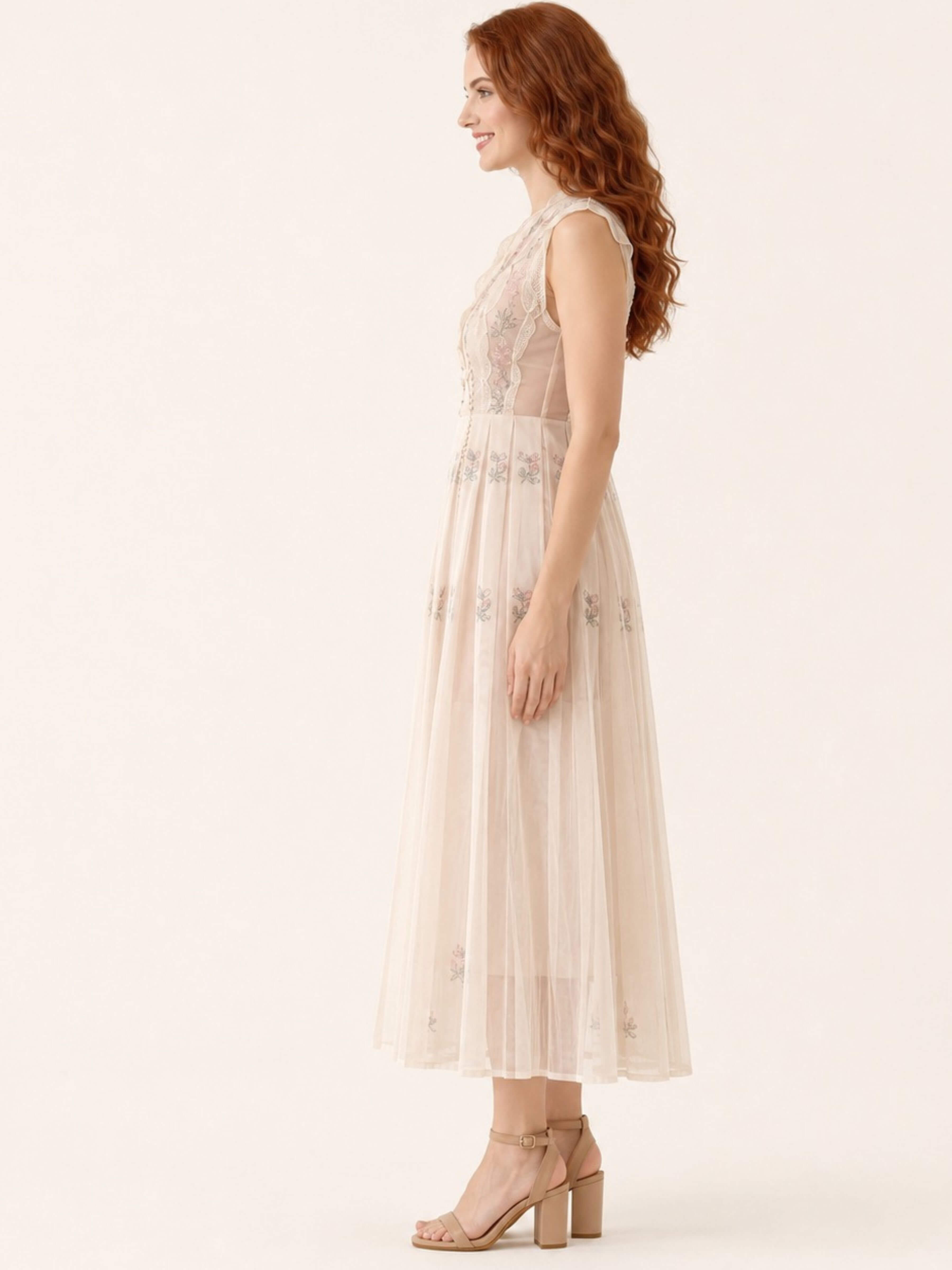 A woman in a sleeveless, floor-length cream tulle dress with floral embroidery and sheer lace bodice, paired with beige block-heel sandals, shown in profile against a warm off-white studio background.