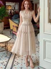 A woman wearing a sleeveless beige midi dress with floral embroidery and a pleated skirt, standing in a decorated room with patterned tile floors and a white door frame.