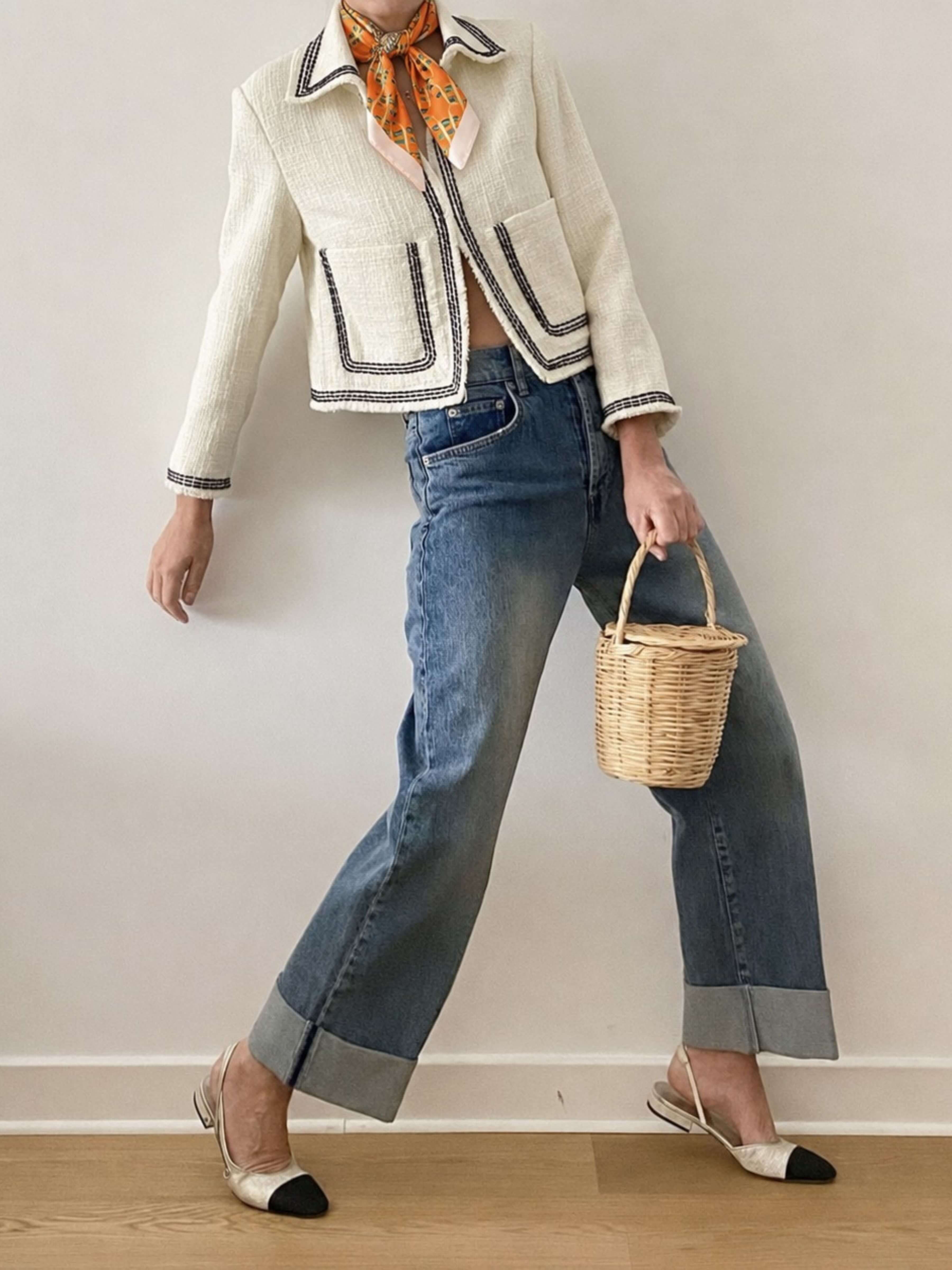 A model is shown from the neck down wearing a cream-colored textured jacket with dark piping, and an orange patterned scarf with blue wide-leg jeans with cuffed hems, holding a small woven basket bag, and wearing white and black pointed-toe flats.