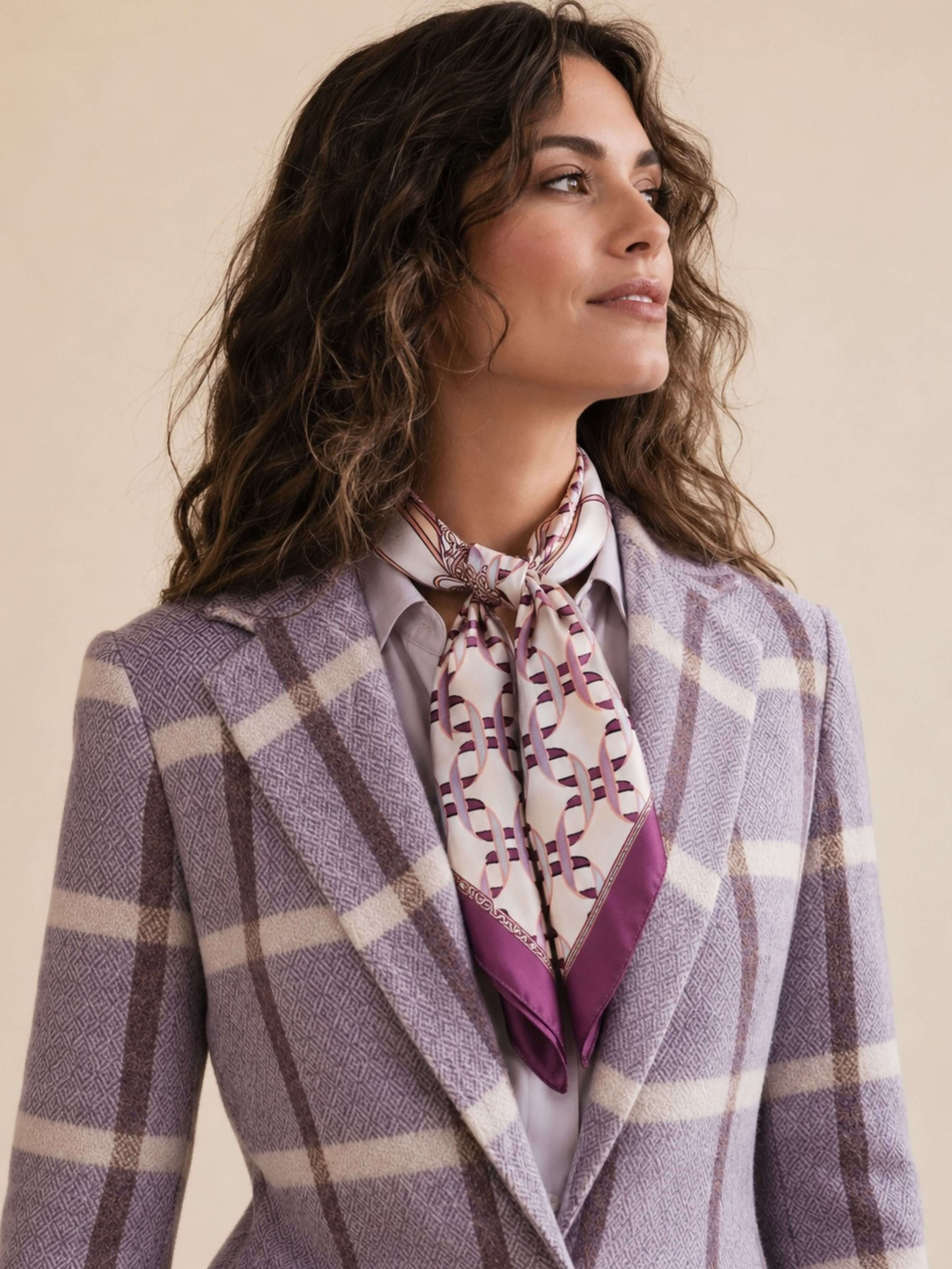 A woman wearing a lavender and beige plaid blazer with a patterned purple and white neck scarf, set against a solid beige background.