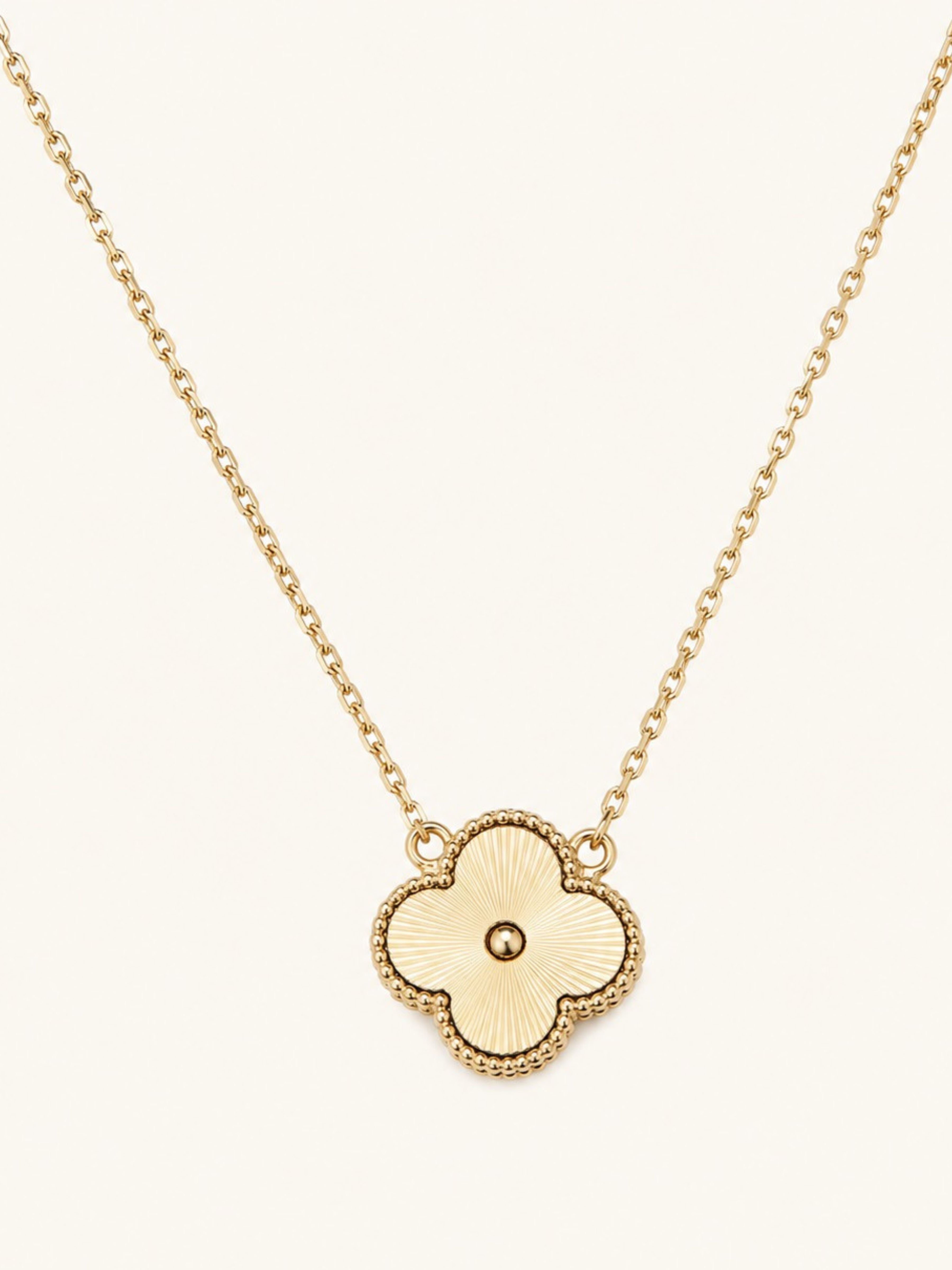 A gold necklace featuring a four-lobed clover pendant with a textured center and a beaded border, suspended from a delicate gold chain against a light cream background.