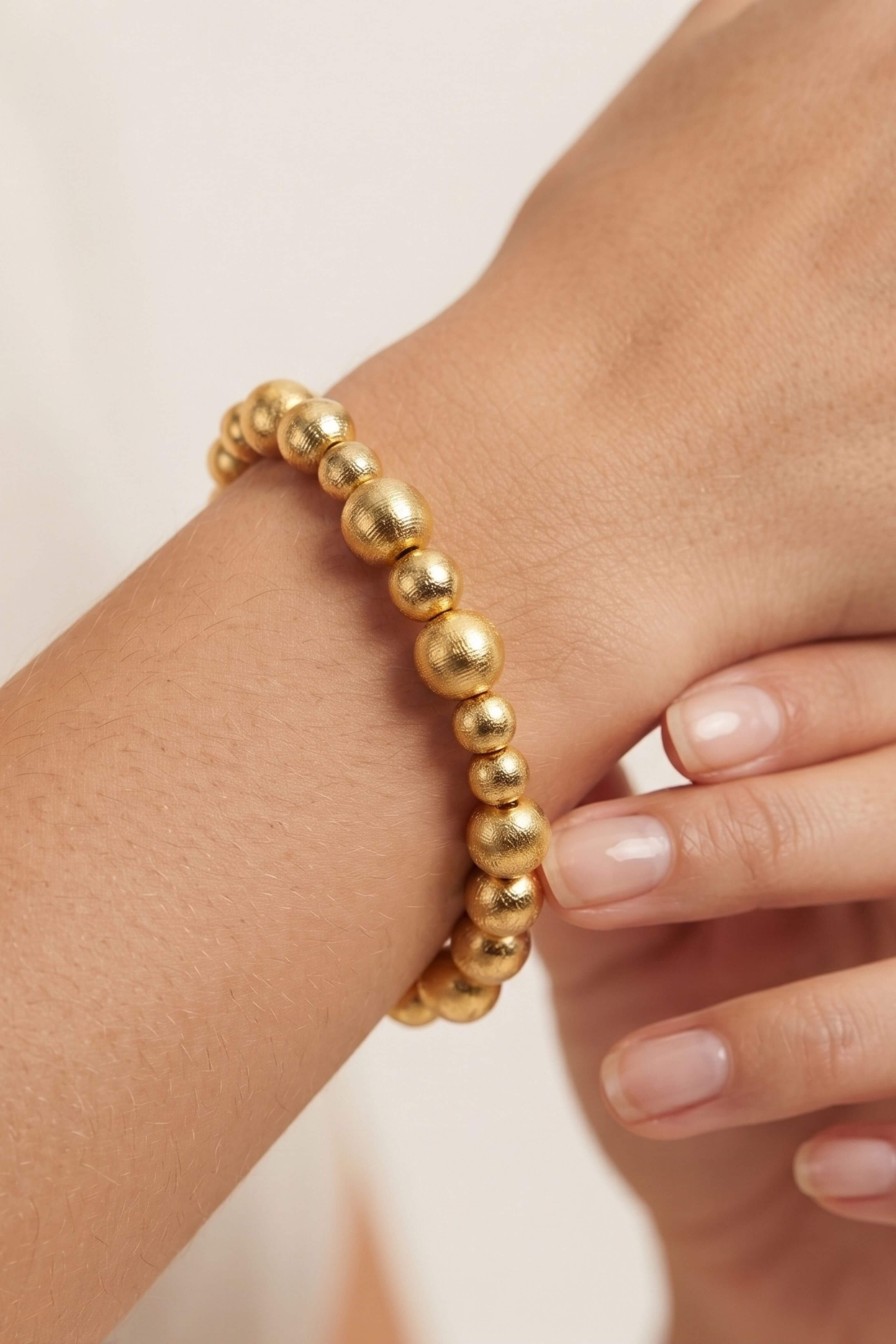 A close-up of a gold beaded stretch bracelet worn on a person's wrist, with fingers gently touching the bracelet against a soft light background.