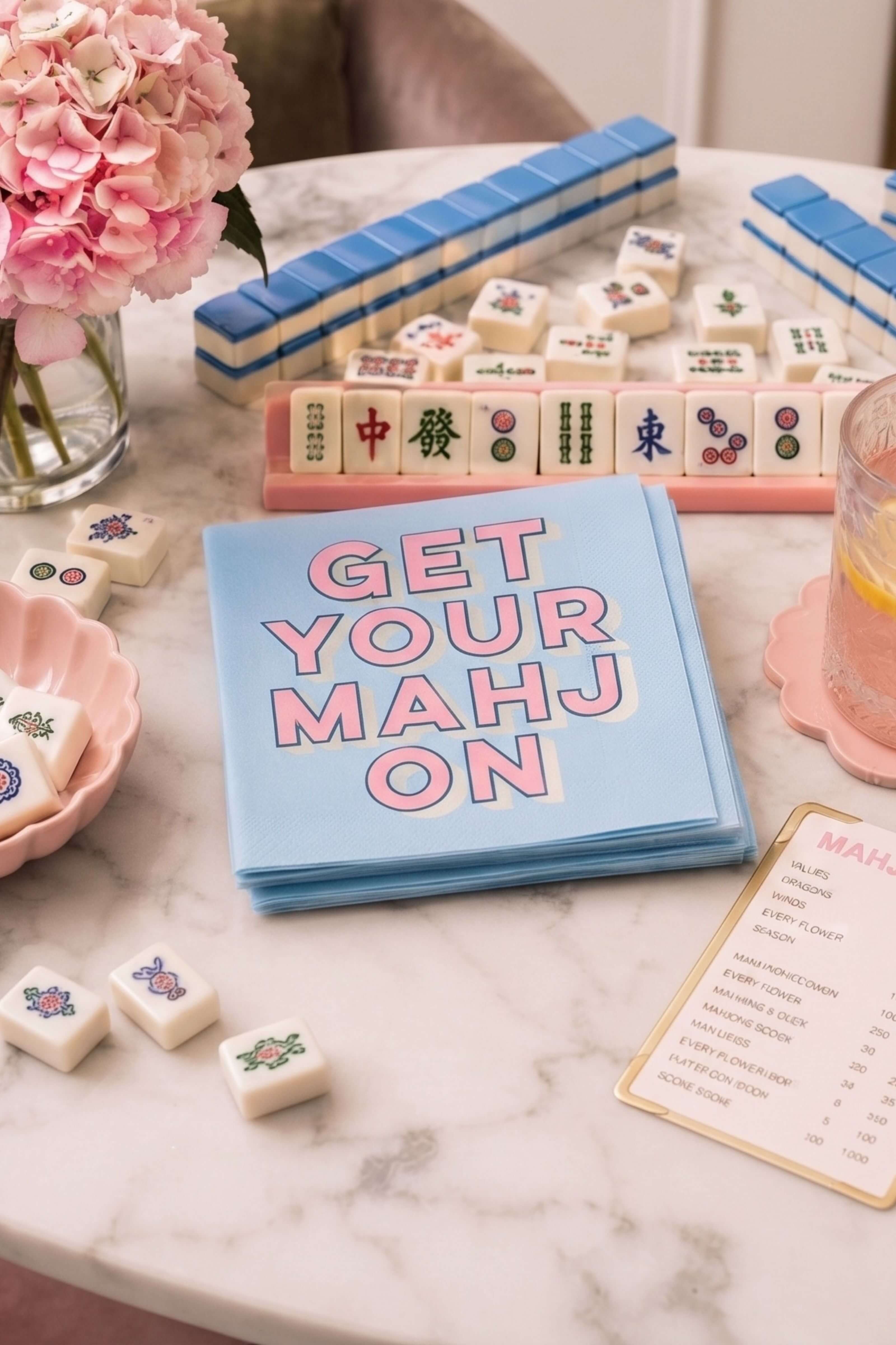 A stack of light blue cards with the phrase 'GET YOUR MAHJON' in pink letters, styled on a marble tabletop with mahjong tiles, pink flowers, and a drink.