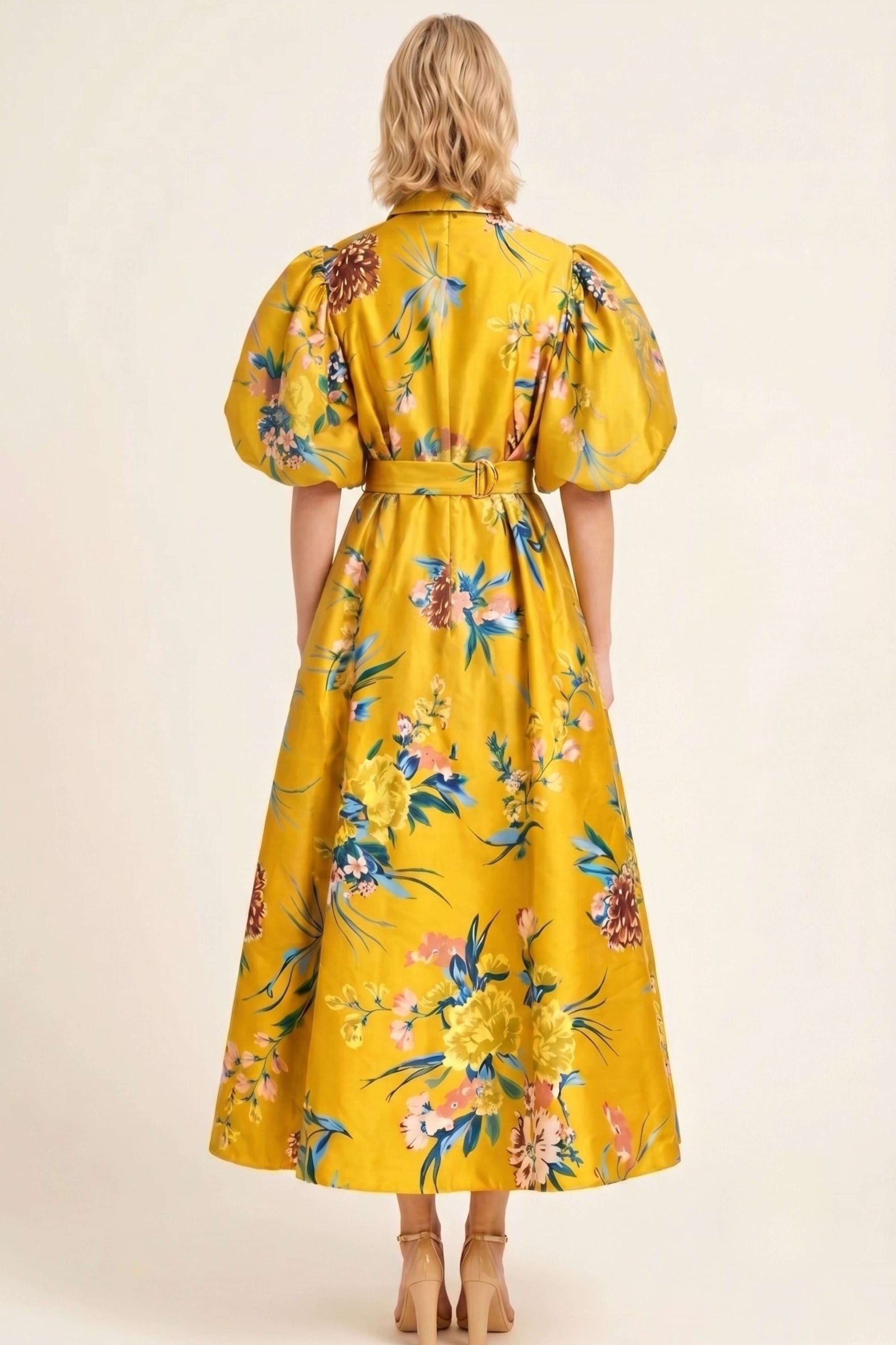 A woman viewed from the back wearing a bright yellow midi dress with a colorful floral print, puff sleeves, and a matching belt, set against a plain beige studio background.
