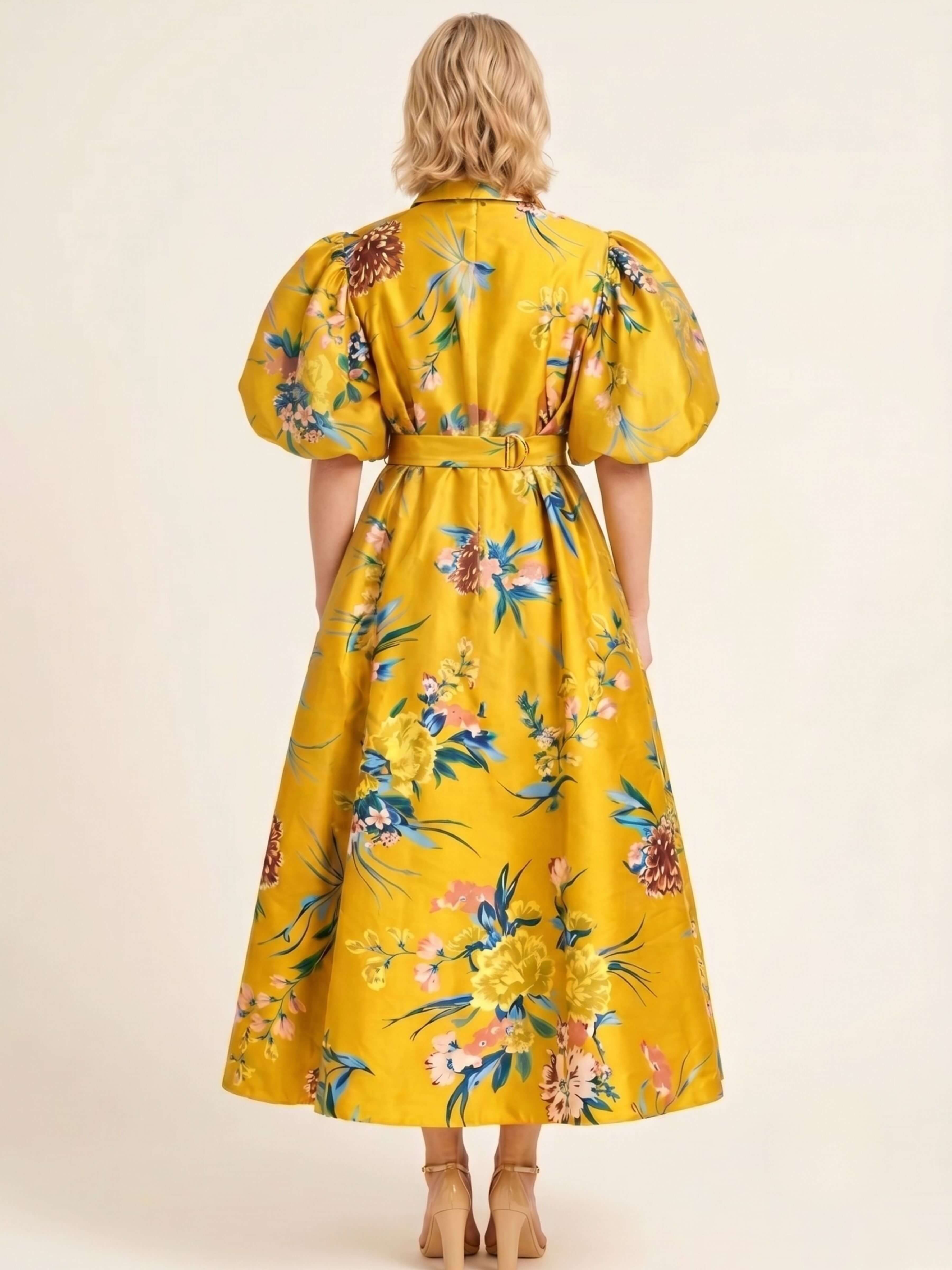 A woman viewed from the back wearing a bright yellow midi dress with a colorful floral print, puff sleeves, and a matching belt, set against a plain beige studio background.