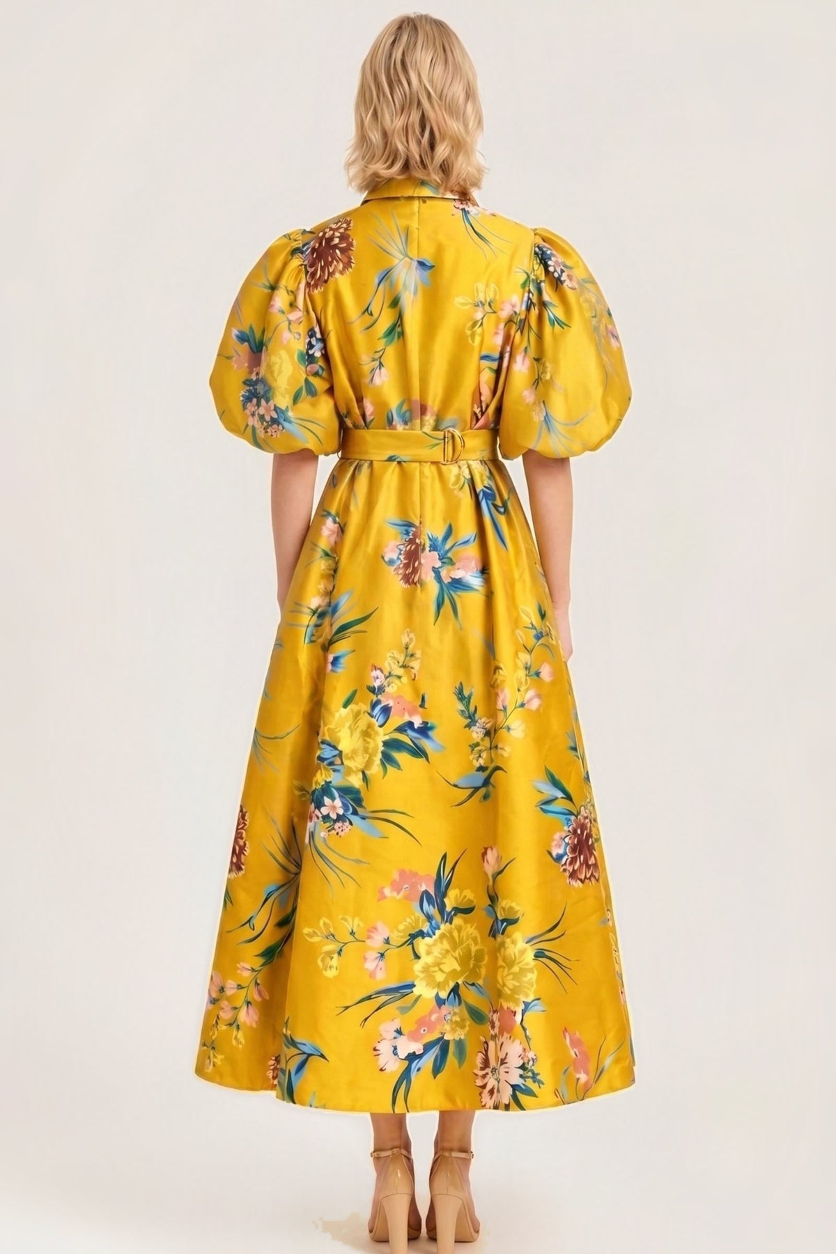 A woman viewed from the back wearing a bright yellow midi dress with a colorful floral print, puff sleeves, and a matching belt, set against a plain beige studio background.