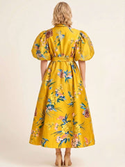 A woman viewed from the back wearing a bright yellow midi dress with a colorful floral print, puff sleeves, and a matching belt, set against a plain beige studio background.
