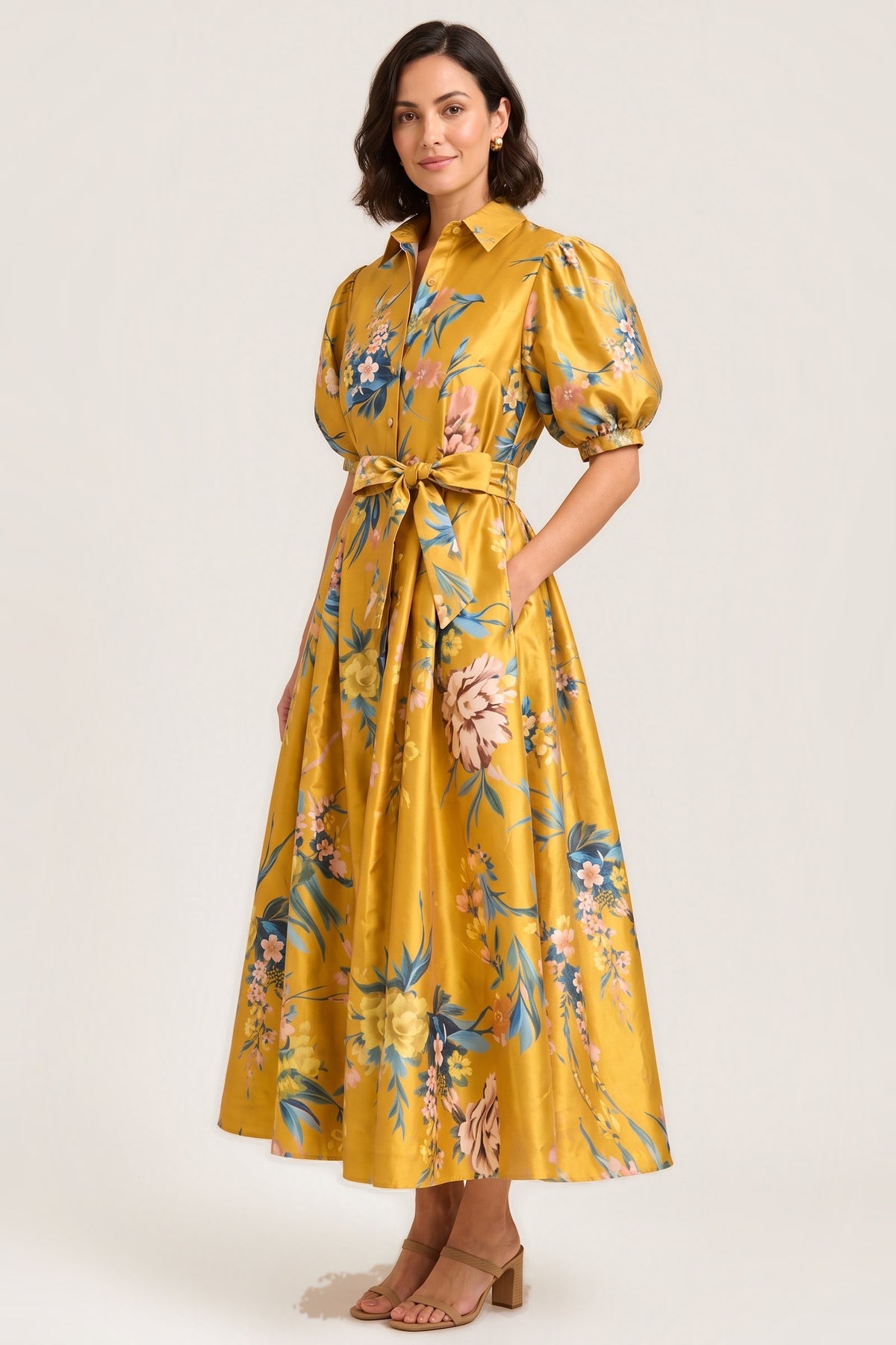 A woman standing in the Golden Garden puff sleeve maxi dress, golden-yellow taffeta with blue and pink painterly florals, puff sleeves and a bow tie waist, one hand in the pocket, on a warm neutral studio background.