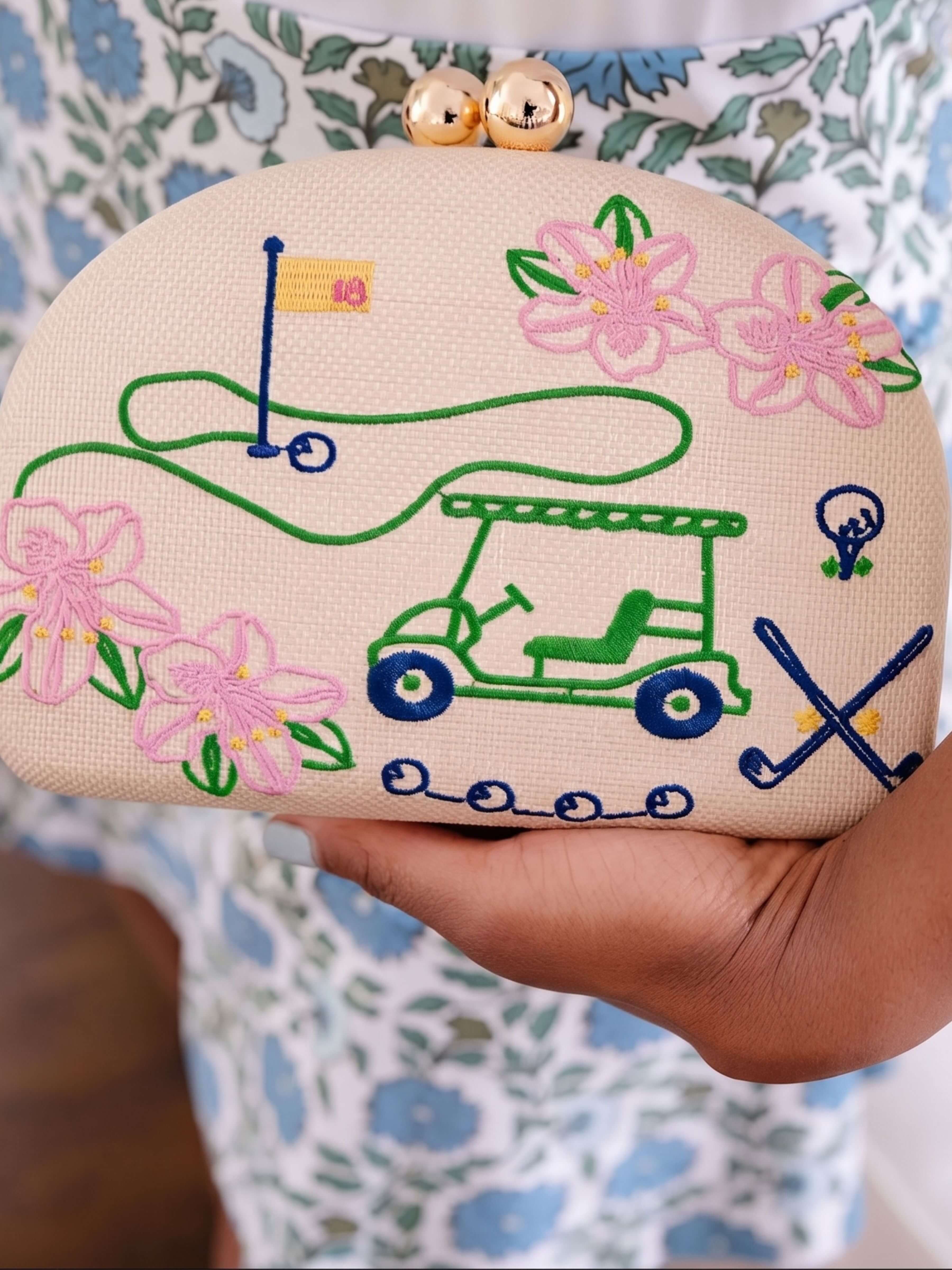 A close-up of a hand holding a beige clutch bag decorated with colorful embroidery of a golf course, a golf cart, pink flowers, and blue clubs. The clutch has a gold-tone clasp closure with two round bells, and the background shows a patterned floral dress.