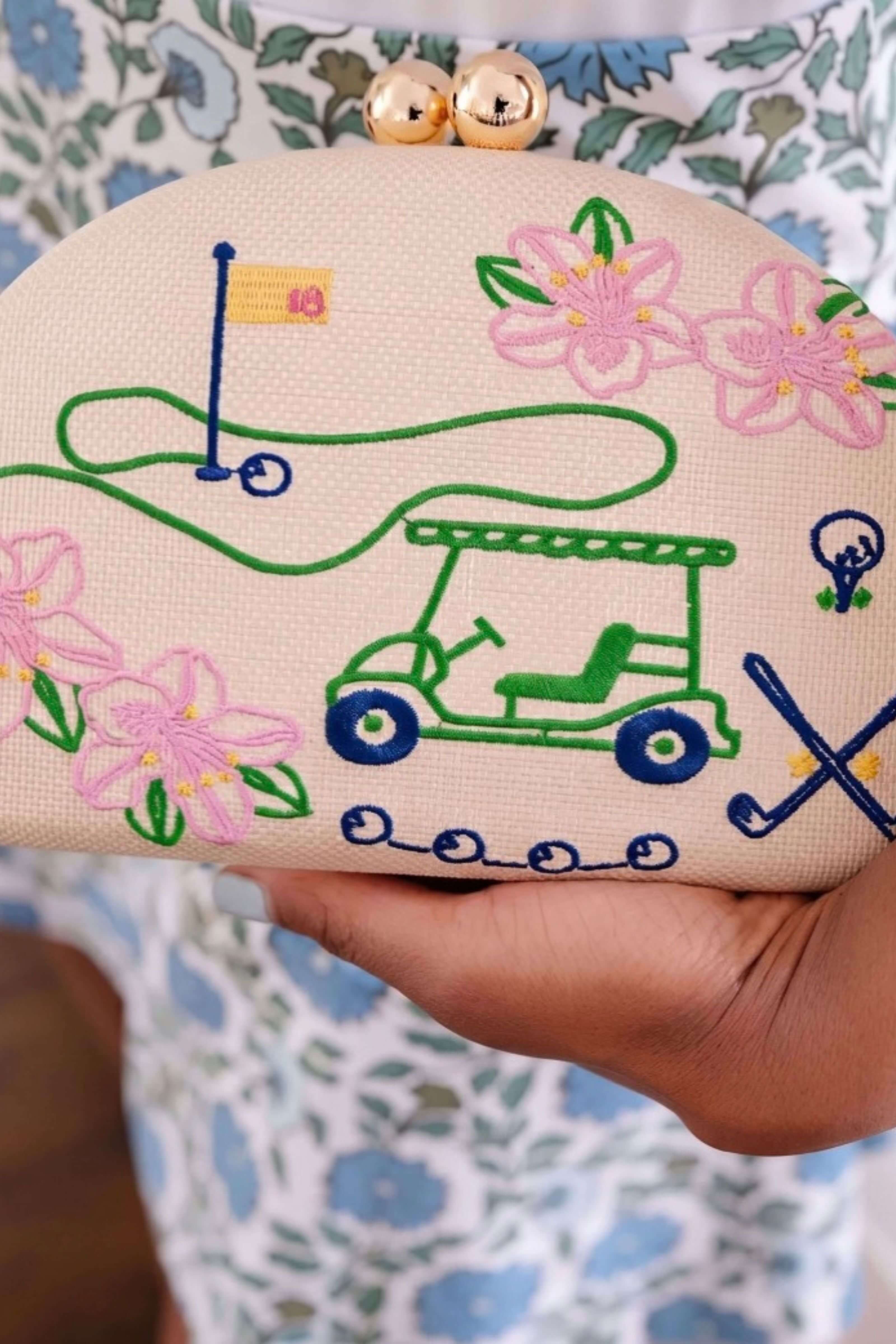 A close-up of a hand holding a beige clutch bag with a golf course and floral design. The clutch features embroidered green and blue details, pink flowers, a golf cart, and a flag, with two gold bell charms at the top.