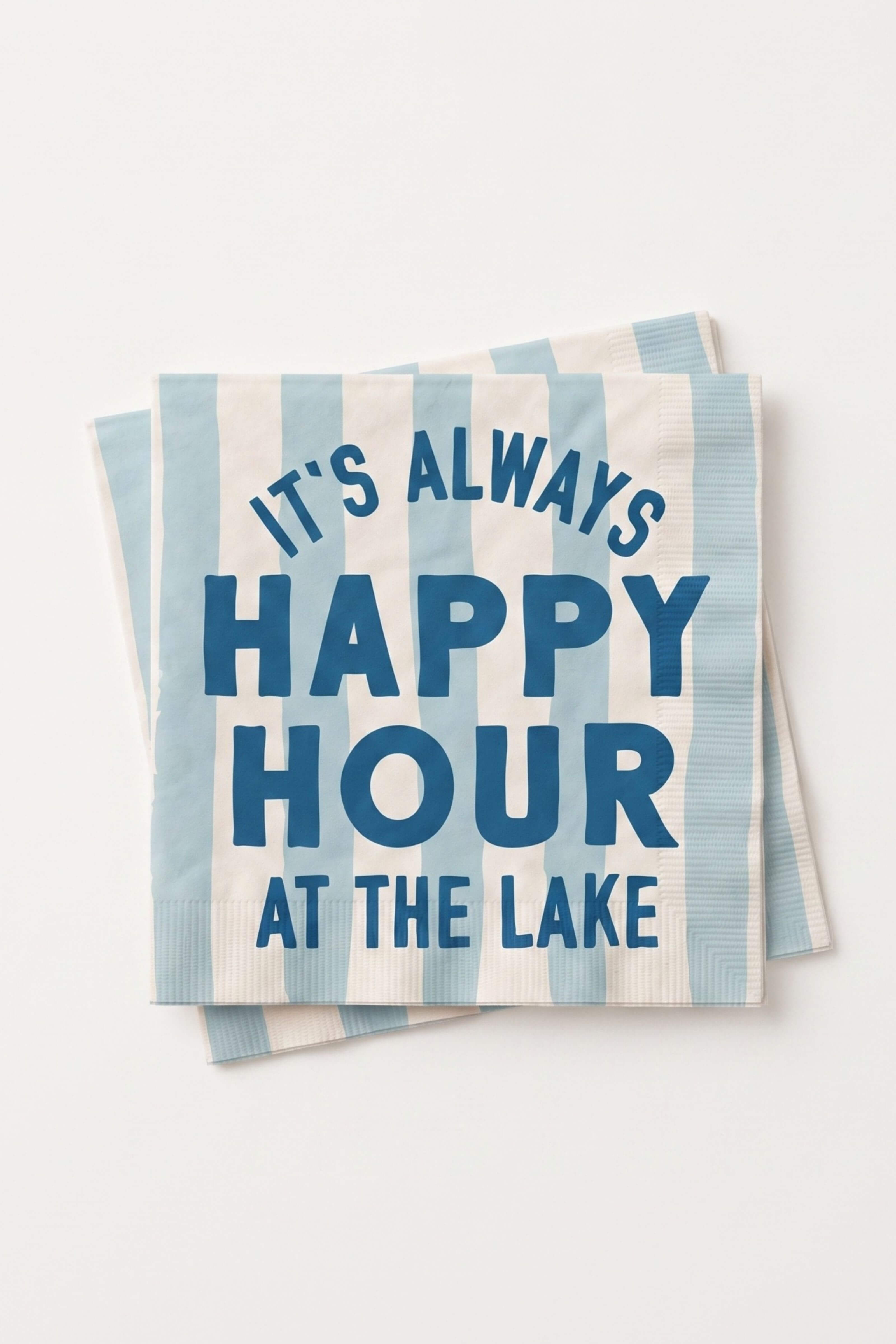 A stack of square paper napkins with light blue and white vertical stripes, featuring the text 'IT'S ALWAYS HAPPY HOUR AT THE LAKE' in dark blue lettering, set against a plain off-white background.