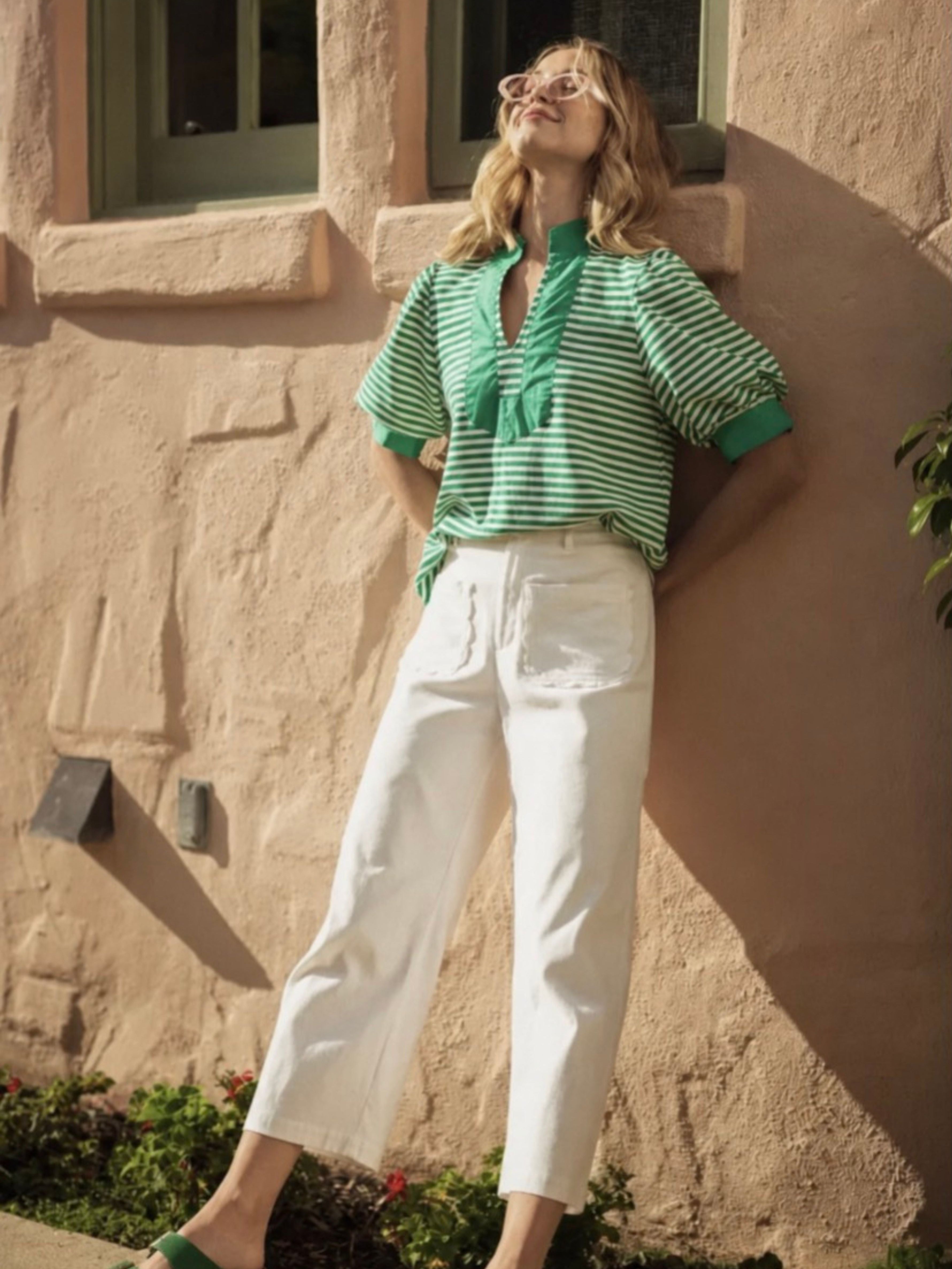 A woman wearing a green and white striped short-sleeve blouse with white cropped pants, standing outdoors against a beige stucco wall.