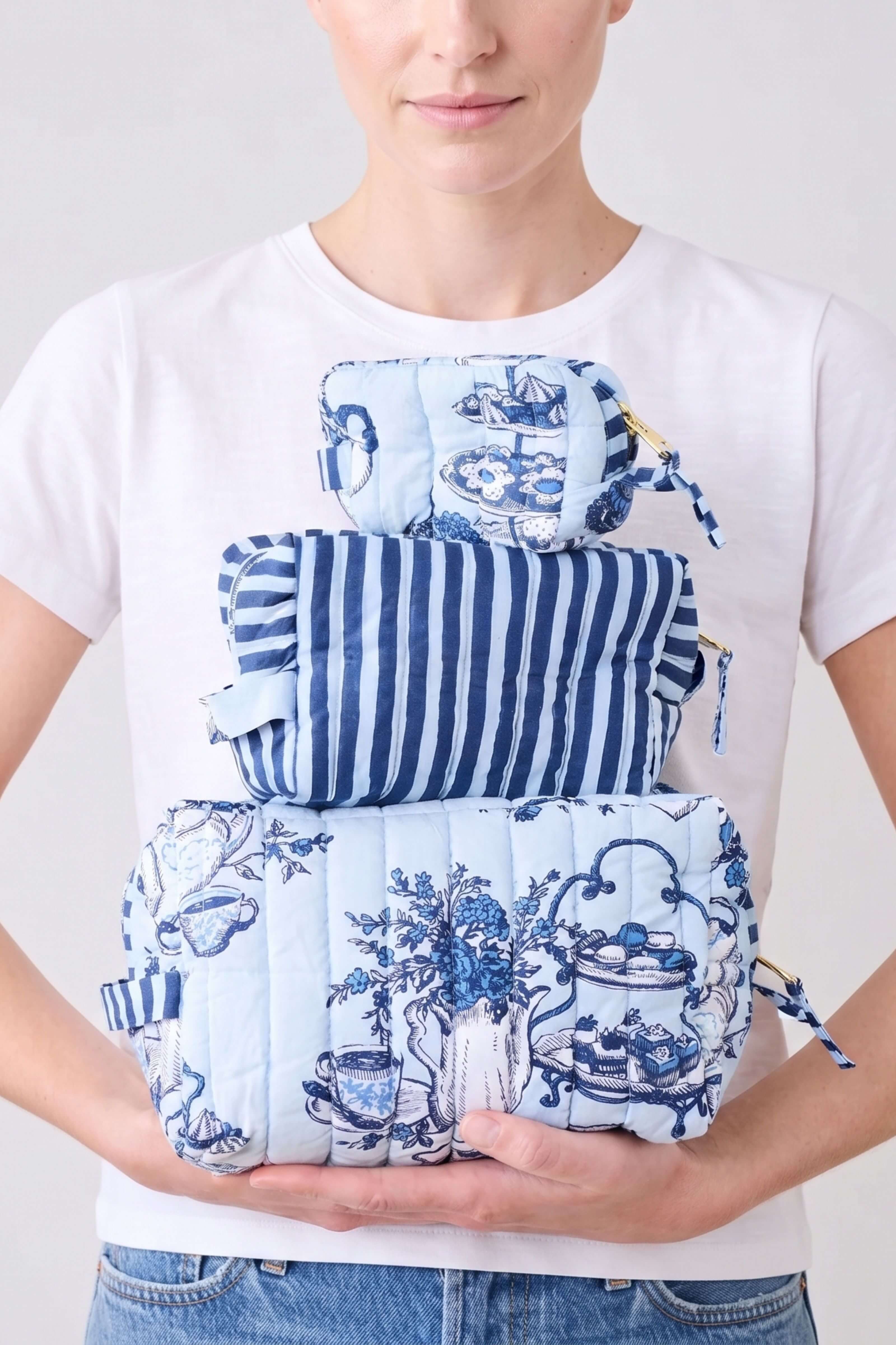 A model wearing a white t-shirt holds three blue and white patterned fabric pouches stacked vertically. The top pouch has a toile-style floral print, the middle pouch has vertical blue and white stripes, and the bottom pouch has a quilted blue and white toile pattern.