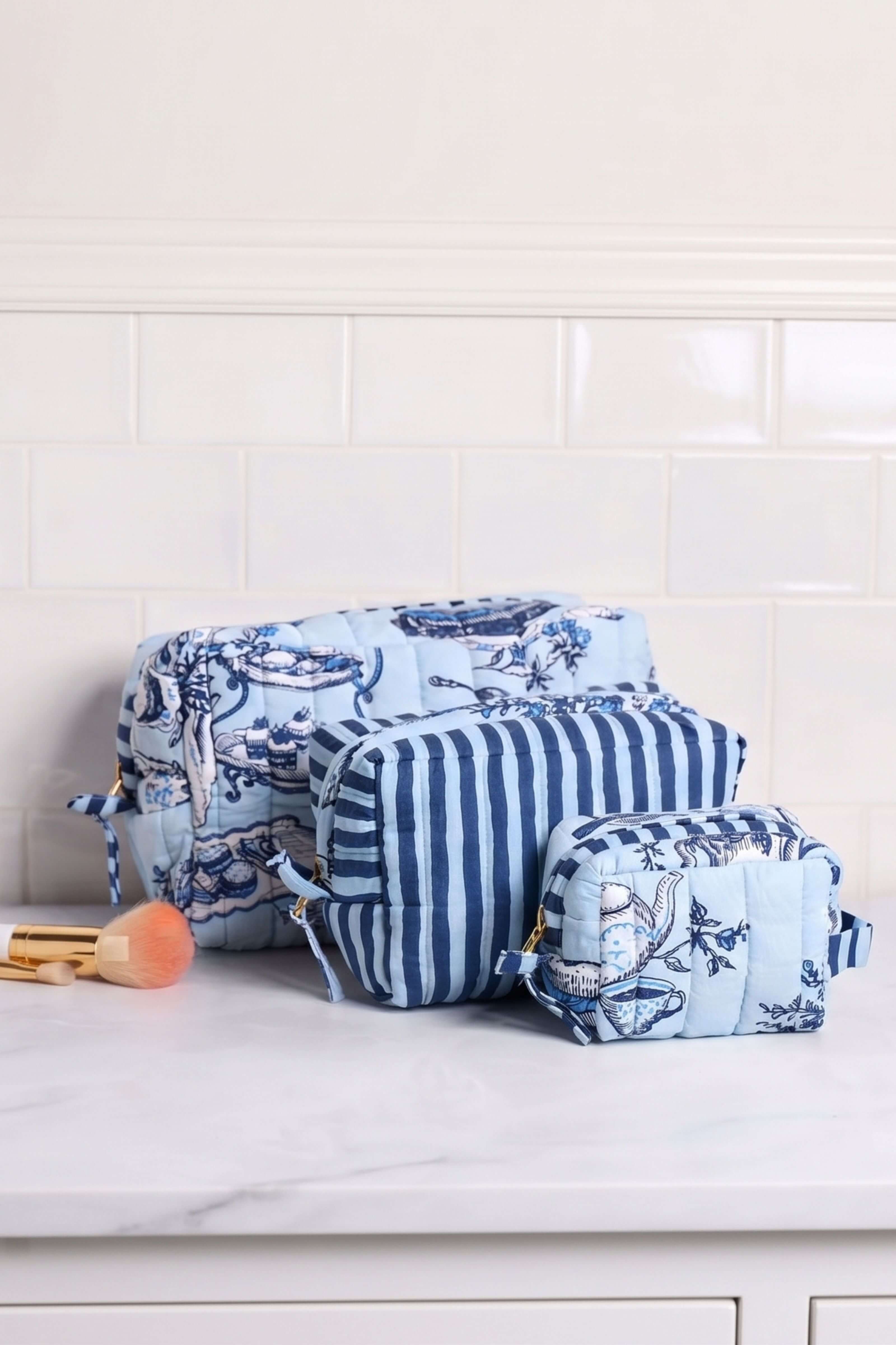 Three blue and white patterned makeup bags arranged on a white marble countertop against a white subway tile wall. One bag features a blue and white floral toile-style print, while the other two feature vertical blue and white stripes.