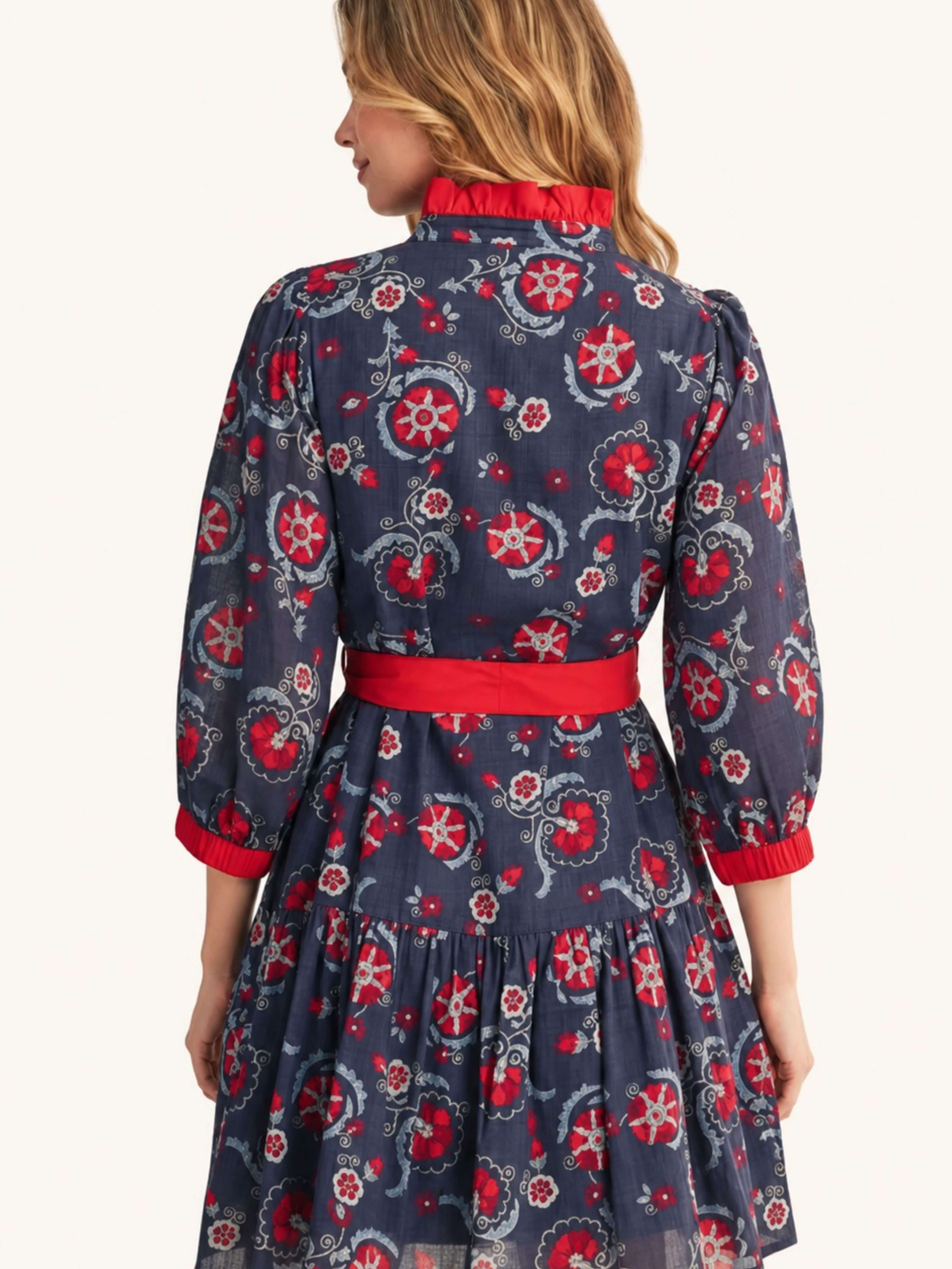 A woman shown from the back wearing a navy blue midi dress with a red and white floral pattern, a red waist belt, and a red ruffled collar, set against a plain off-white background.