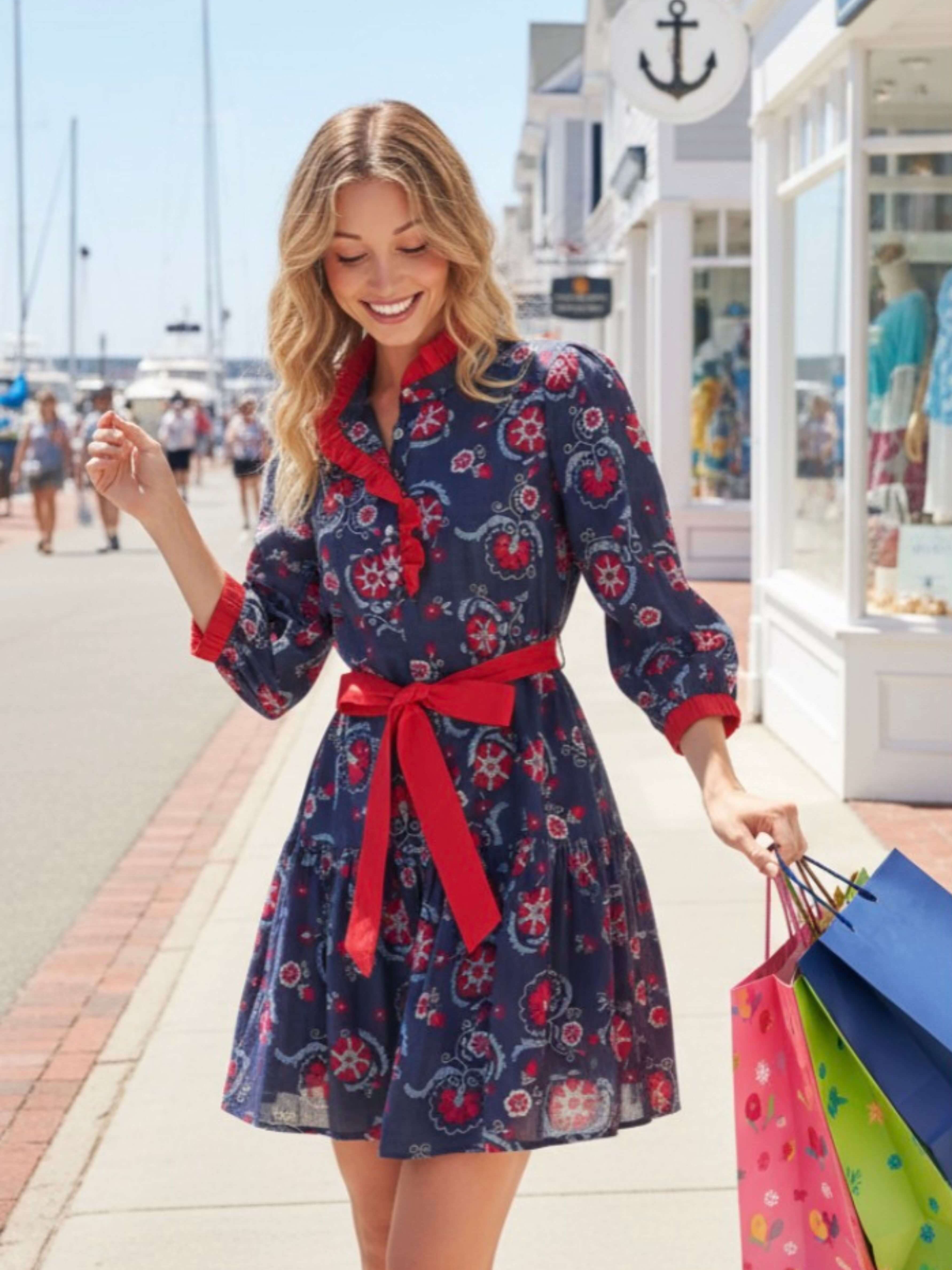 A woman walking on a sunny seaside promenade wearing a navy blue mini dress with a red and pink floral pattern, a red ruffled collar, and a red waist tie. She is holding colorful shopping bags.