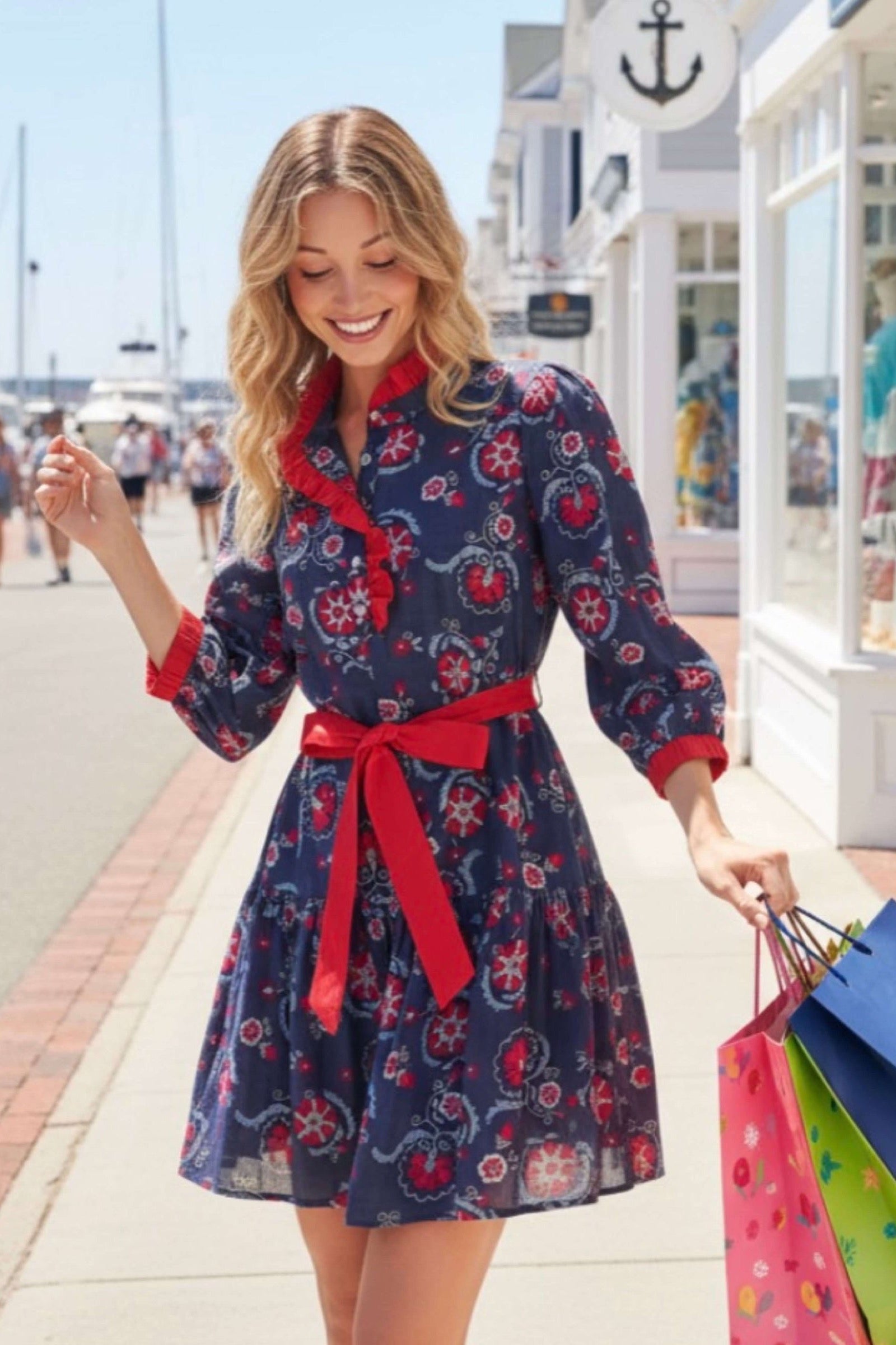 A woman walking on a sunny seaside promenade wearing a navy blue mini dress with a red and pink floral pattern, a red ruffled collar, and a red waist tie. She is holding colorful shopping bags.