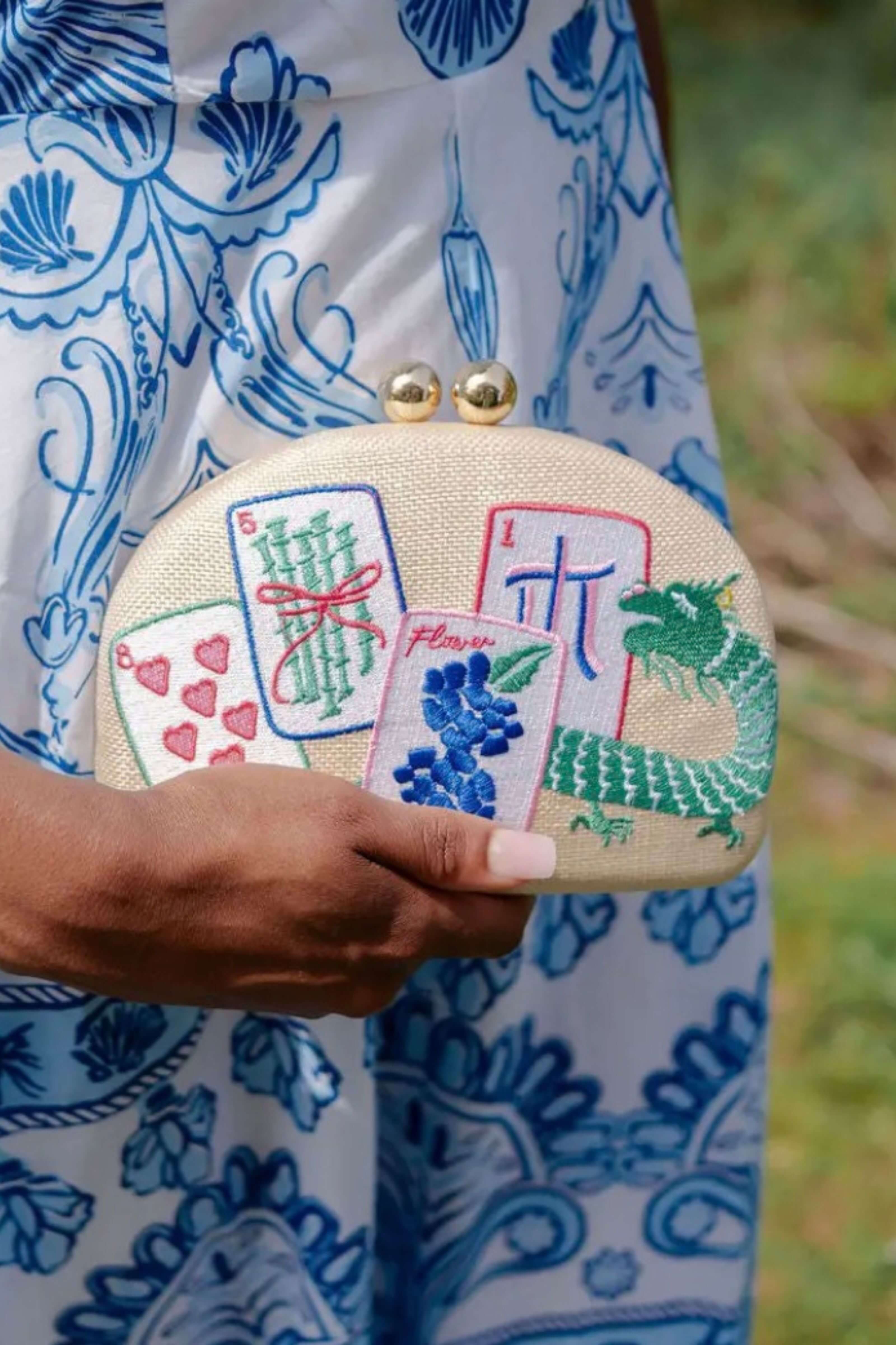 A close-up of a person holding a cream-colored clutch purse decorated with embroidered mahjong tiles and playing card motifs. The person is wearing a blue and white patterned dress, with a soft-focus green outdoor background.