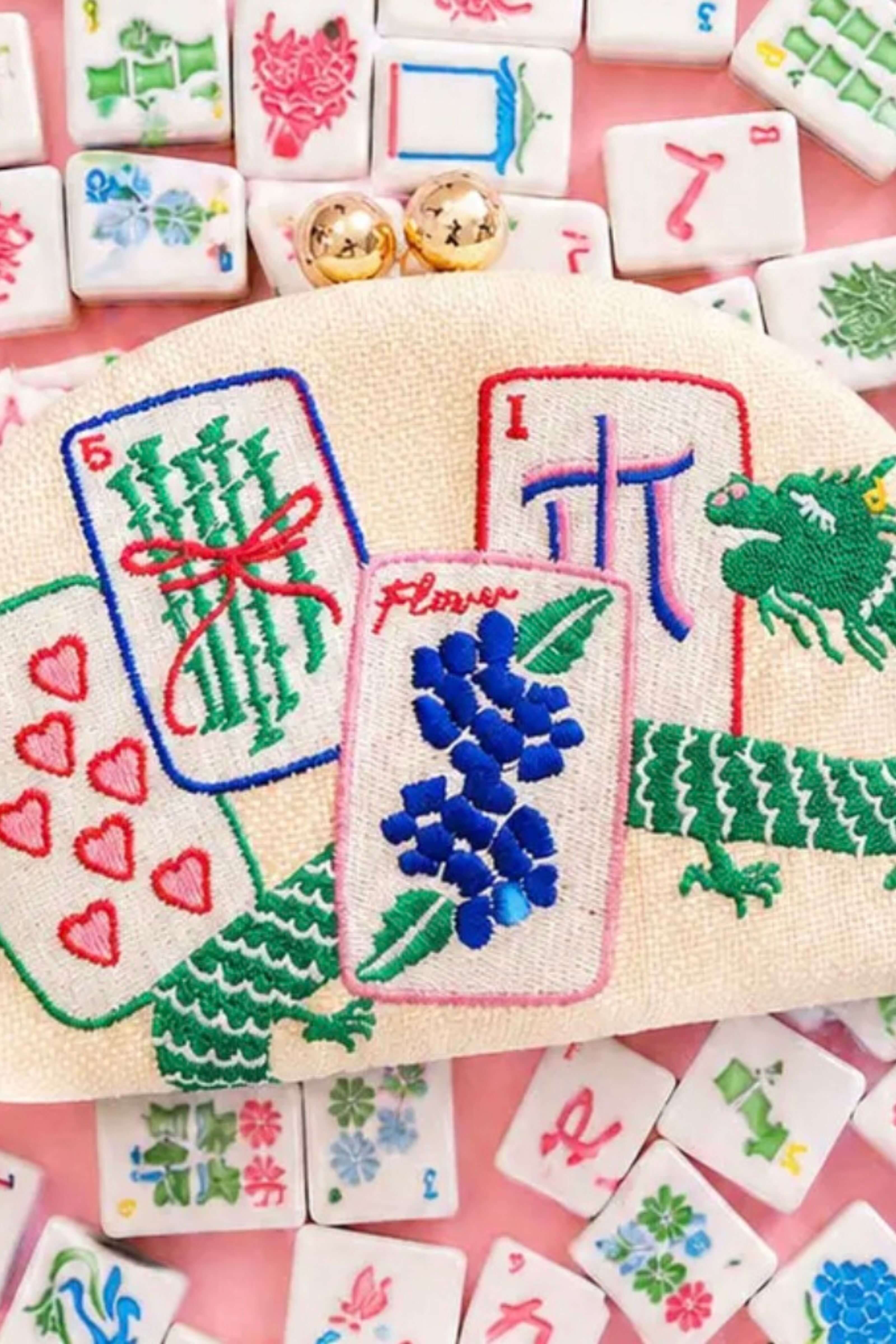 A beige canvas coin purse with colorful embroidered mahjong tile designs, surrounded by scattered white and pink mahjong tiles on a pink background.