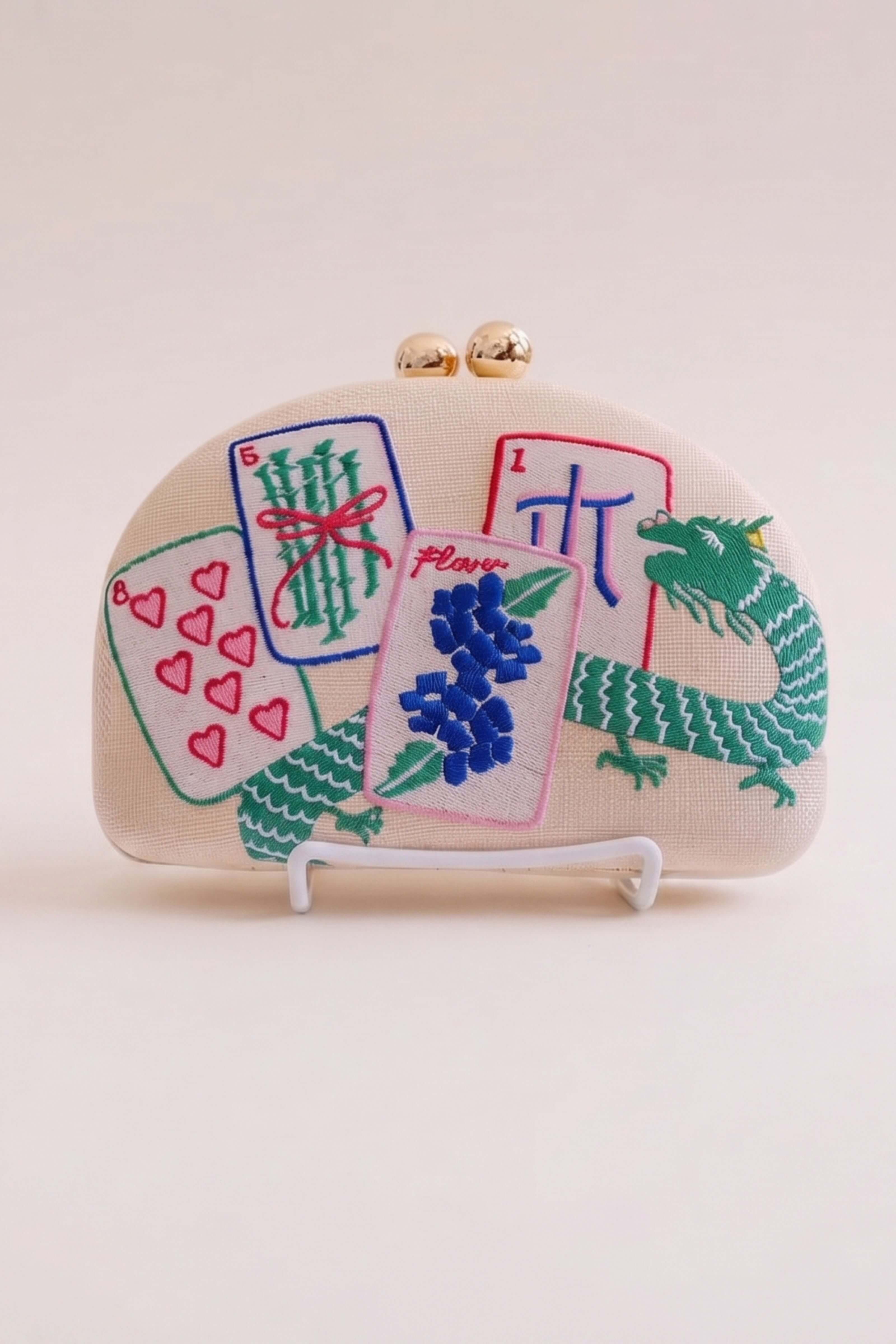 A cream-colored clutch purse with gold-tone metal clasp hardware, decorated with colorful embroidered Chinese mahjong tiles and a green dragon motif, displayed on a small white stand against a light beige background.