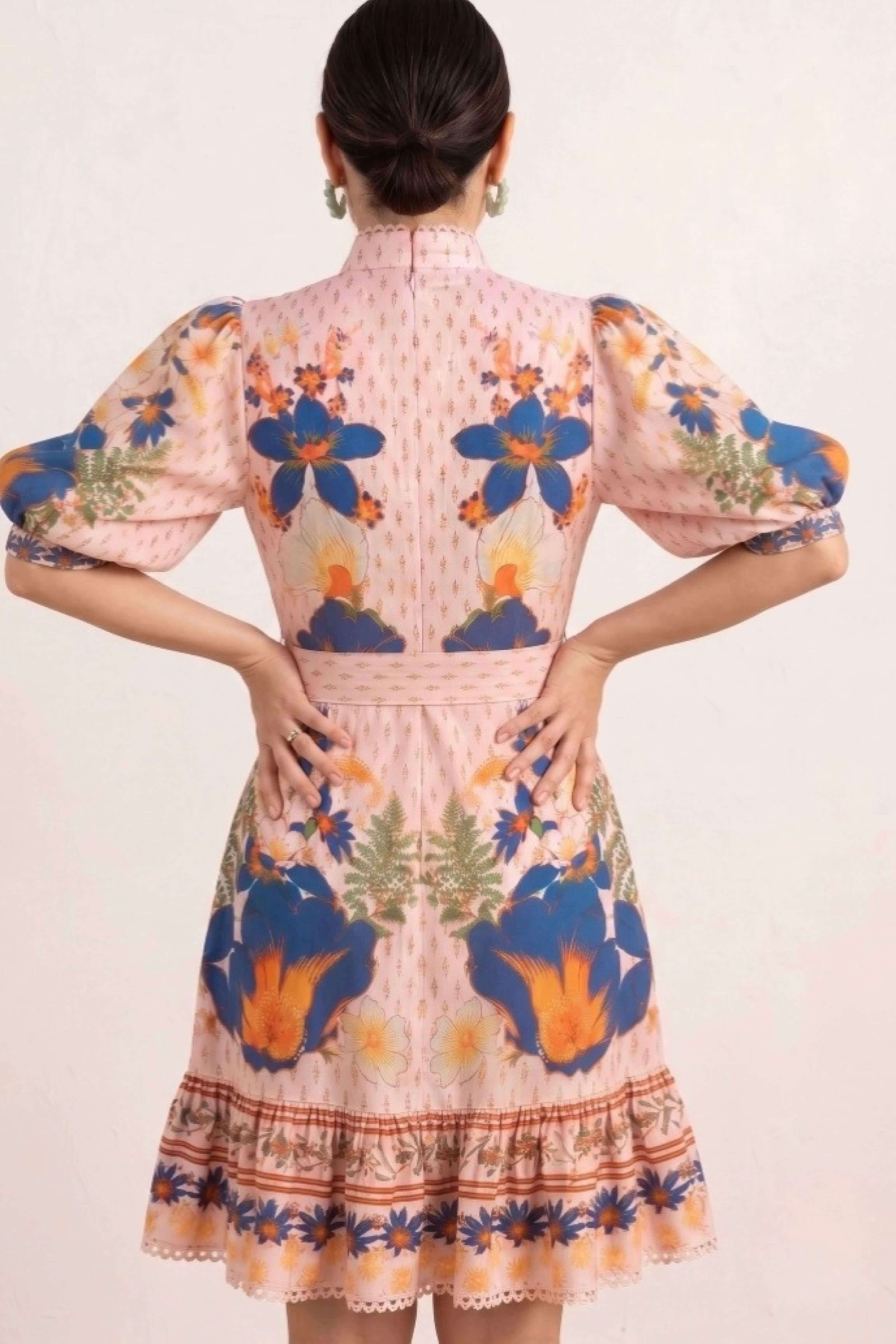 A woman shown from the back wearing a light pink dress with blue, orange, and green floral patterns and puff sleeves, photographed against a plain light background.