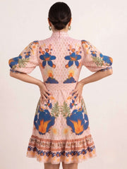 A woman viewed from the back wearing a short pink dress with puff sleeves and a colorful floral print in blue, orange, and green, set against a plain light beige background.