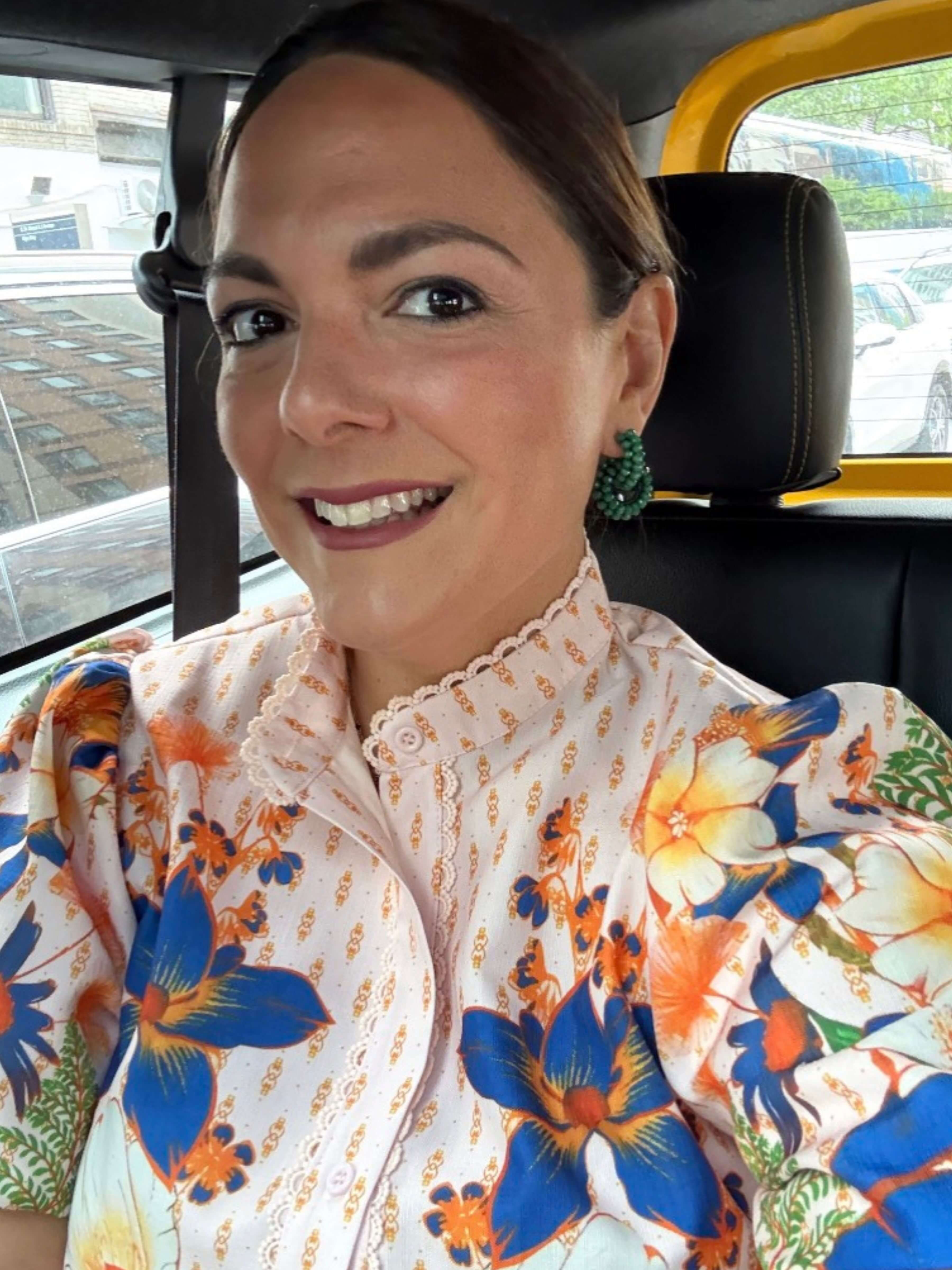 A woman smiling inside a vehicle, wearing a cream-colored blouse with a vibrant blue, orange, and yellow floral print and lace trim.