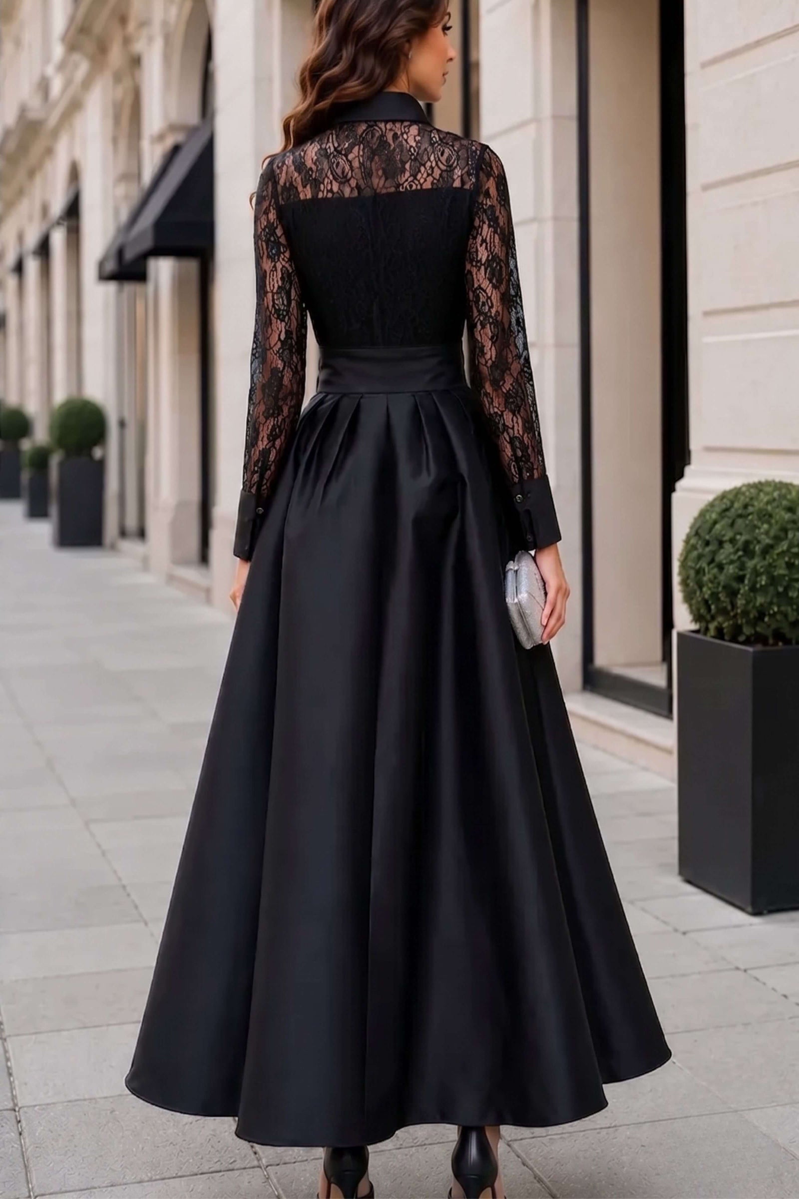 A woman wearing a long black evening gown with sheer black lace long sleeves and a matching black satin waistband, photographed from behind on a city sidewalk.